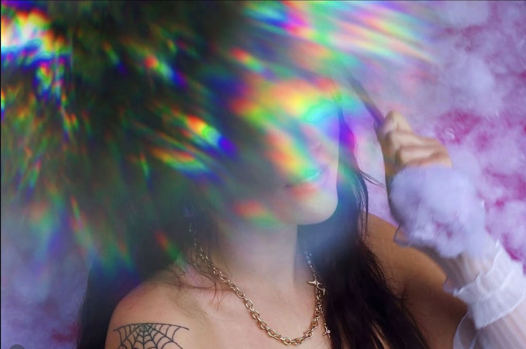 Woman with dark hair and a spider web tattoo on her shoulder, holding a stick and looking at the colorful rainbow-like light across her face and background.
Lifestyle witchy portrait.