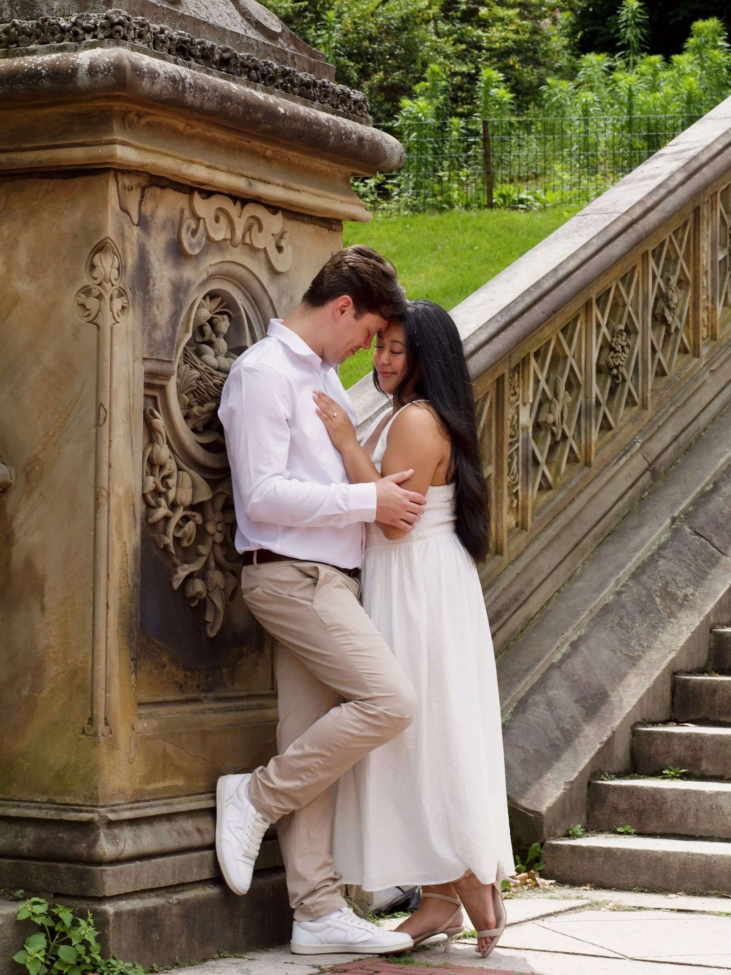 A young man and woman in white and cream clothing standing closely together outdoors, touching foreheads with eyes closed, near an ornate stone structure and a staircase, in a lush green setting.