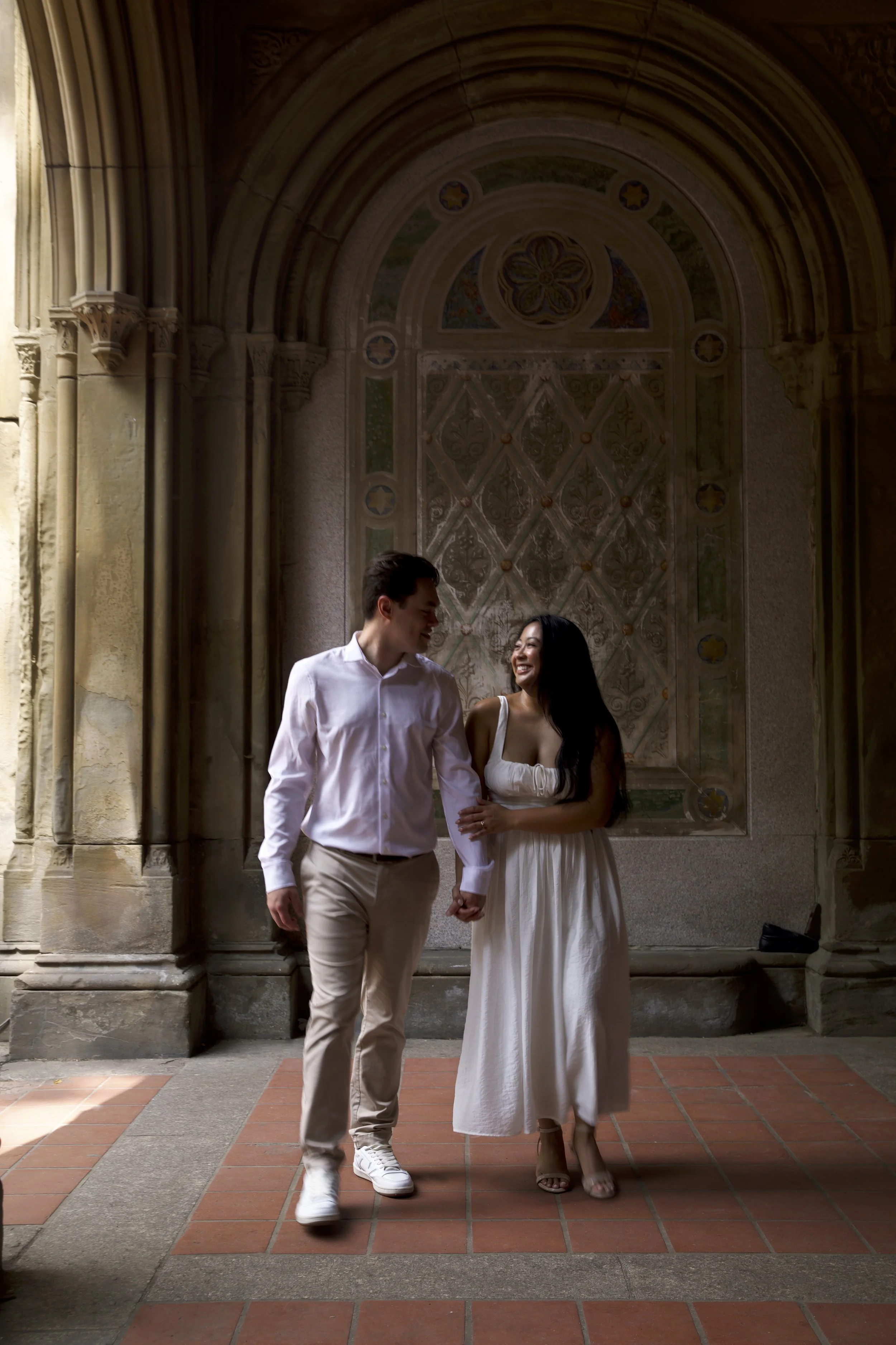 A smiling couple holding hands, walking in a historic architectural setting with arched walls and decorative details. Engagement shoot at Bethesda Terrace in NYC.