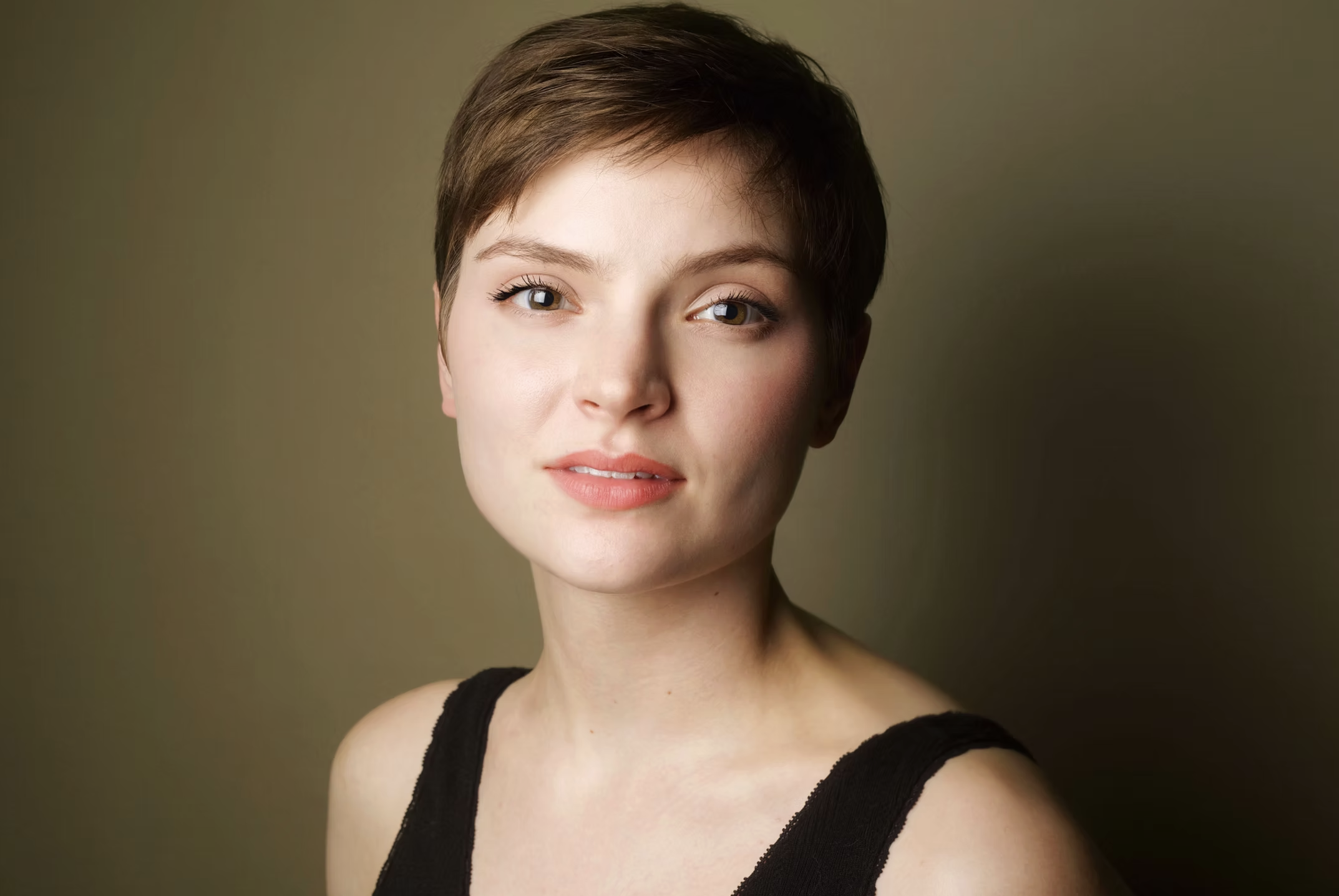 Portrait of a young woman with short brown hair, fair skin, and blue eyes, wearing a black tank top, against a plain green background. Sark and soft headshot.