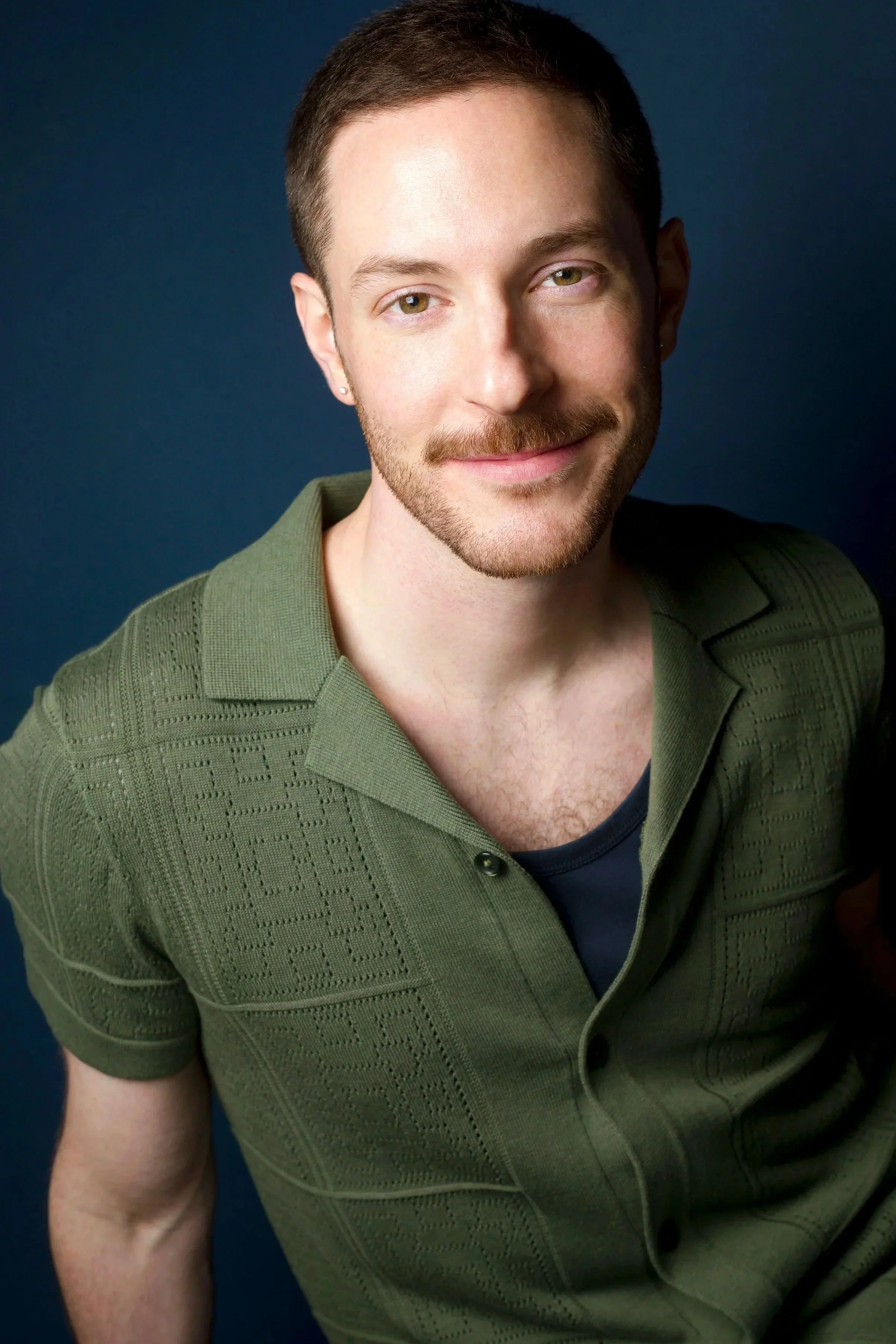 A man with short brown hair, light complexion, green eyes, a light beard and mustache, wearing a green textured polo shirt and smiling, against a dark blue background.