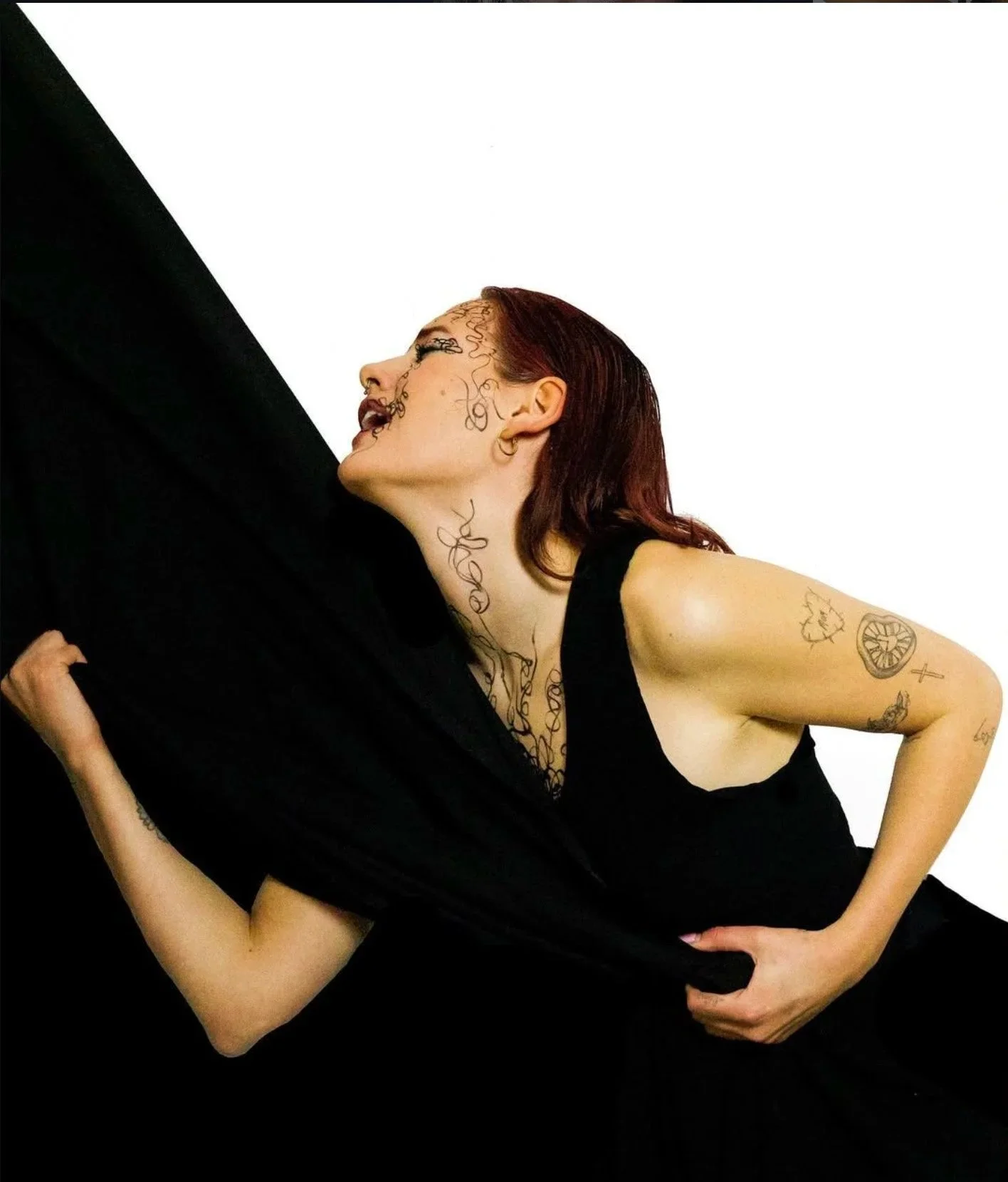 A woman with red hair and tattoos on her neck, arm, and face wearing a black dress, leaning against a large black sheet or fabric, with a white background. Anxiety inspired lifestyle shoot.