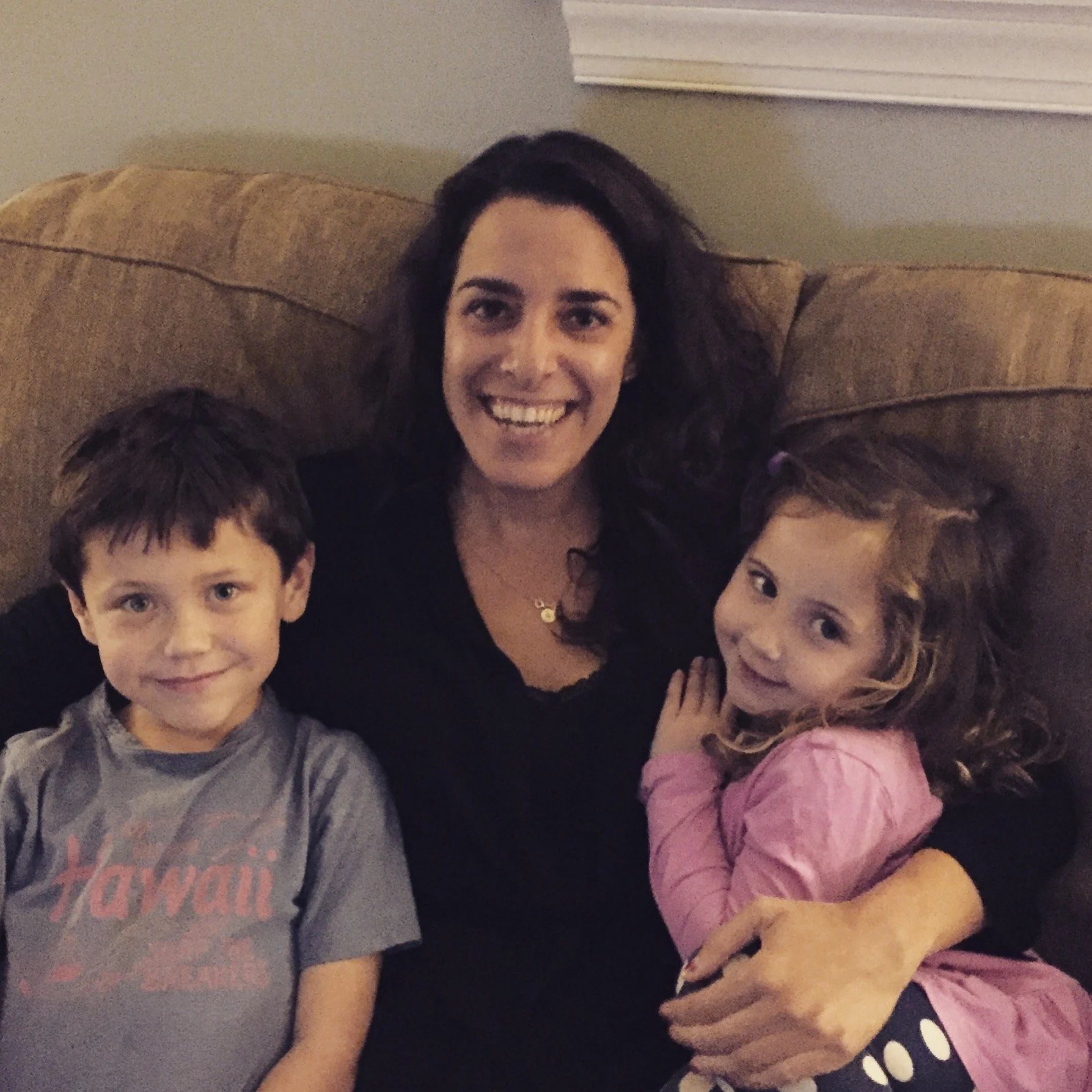A woman sitting on a couch with a young boy and girl, smiling at the camera inside a home.
