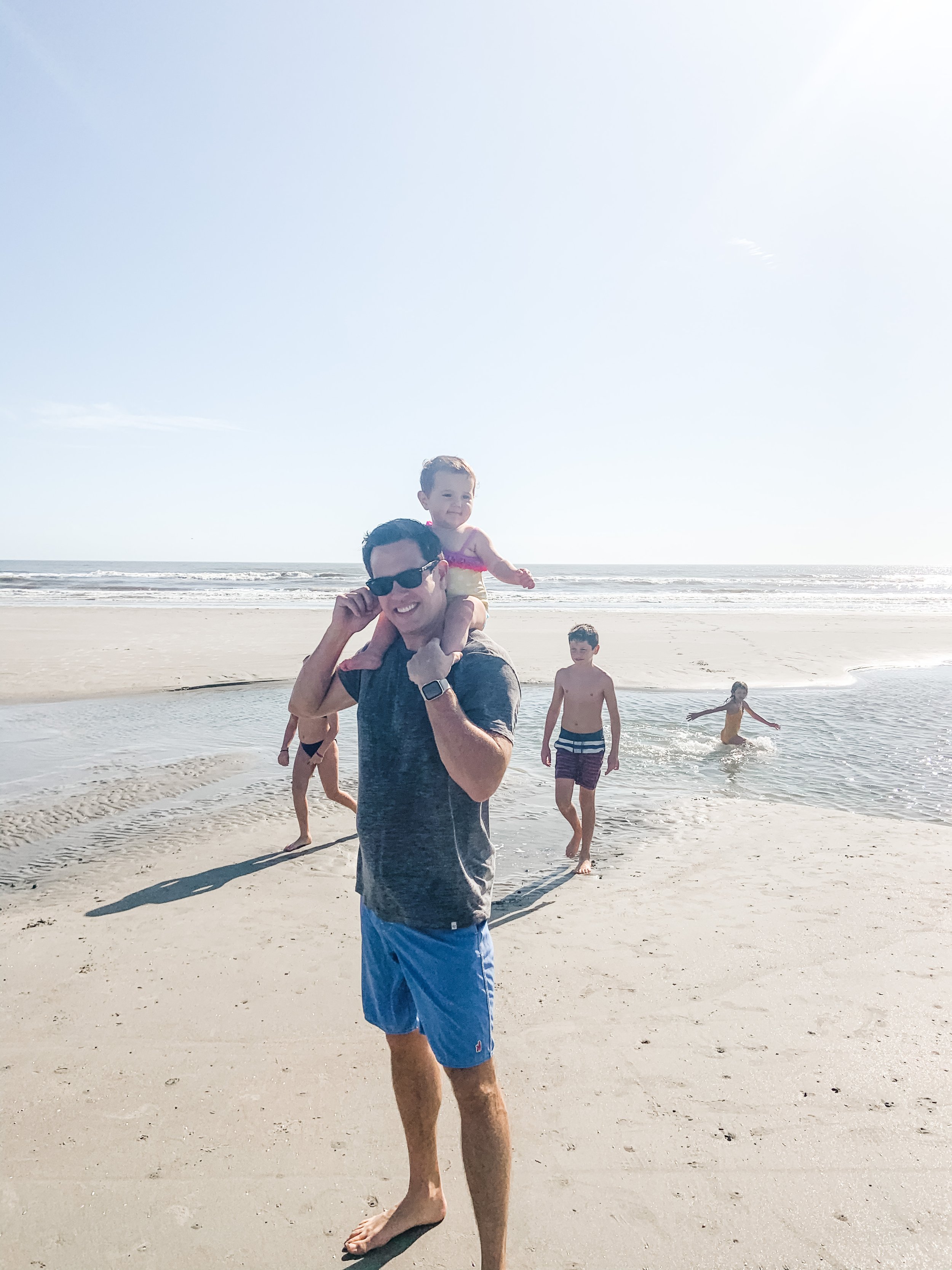 A man wearing sunglasses and blue shorts is carrying a young girl on his shoulders at the beach, with three children playing in the water nearby and a sandy ocean shoreline in the background.