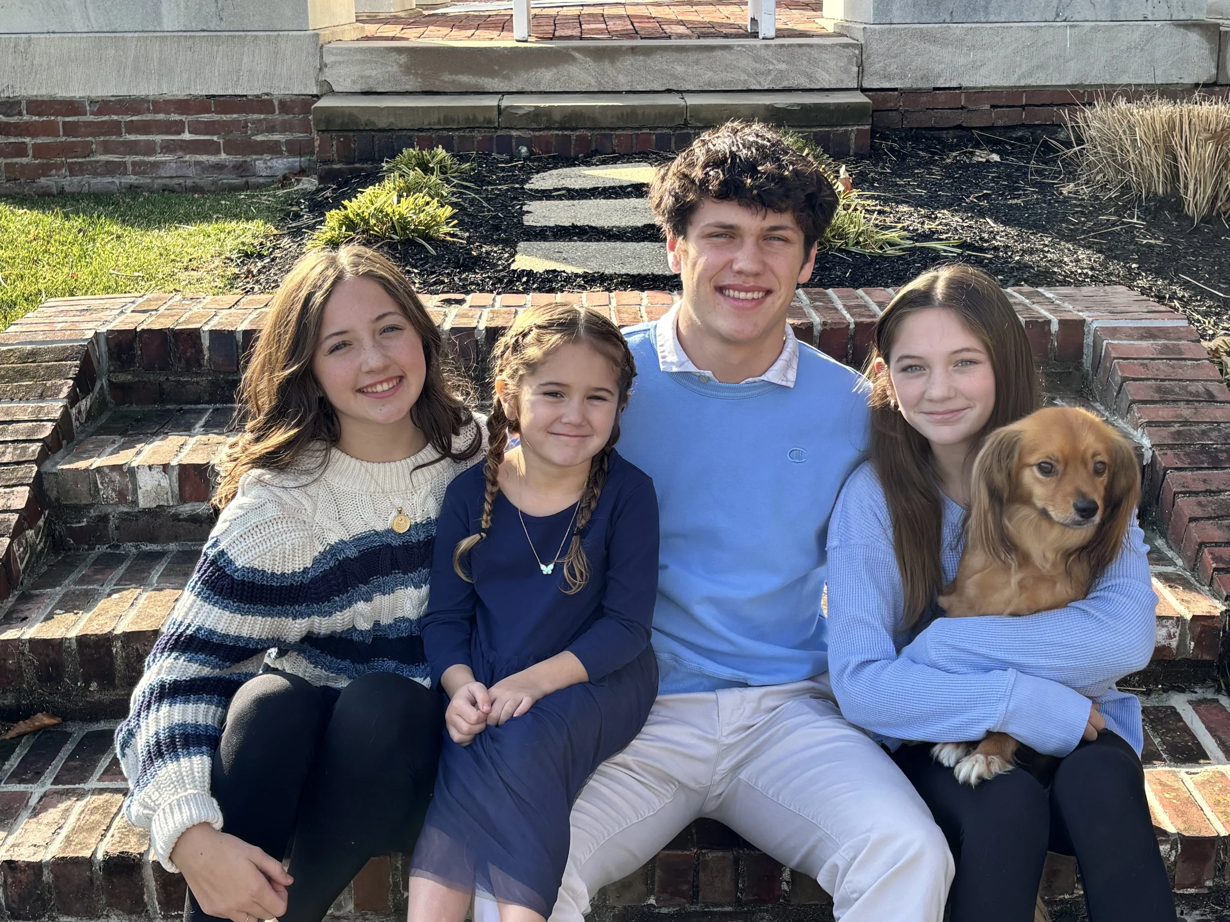 A family of five, including four children and a dog, sitting on brick steps outside a house, smiling at the camera.