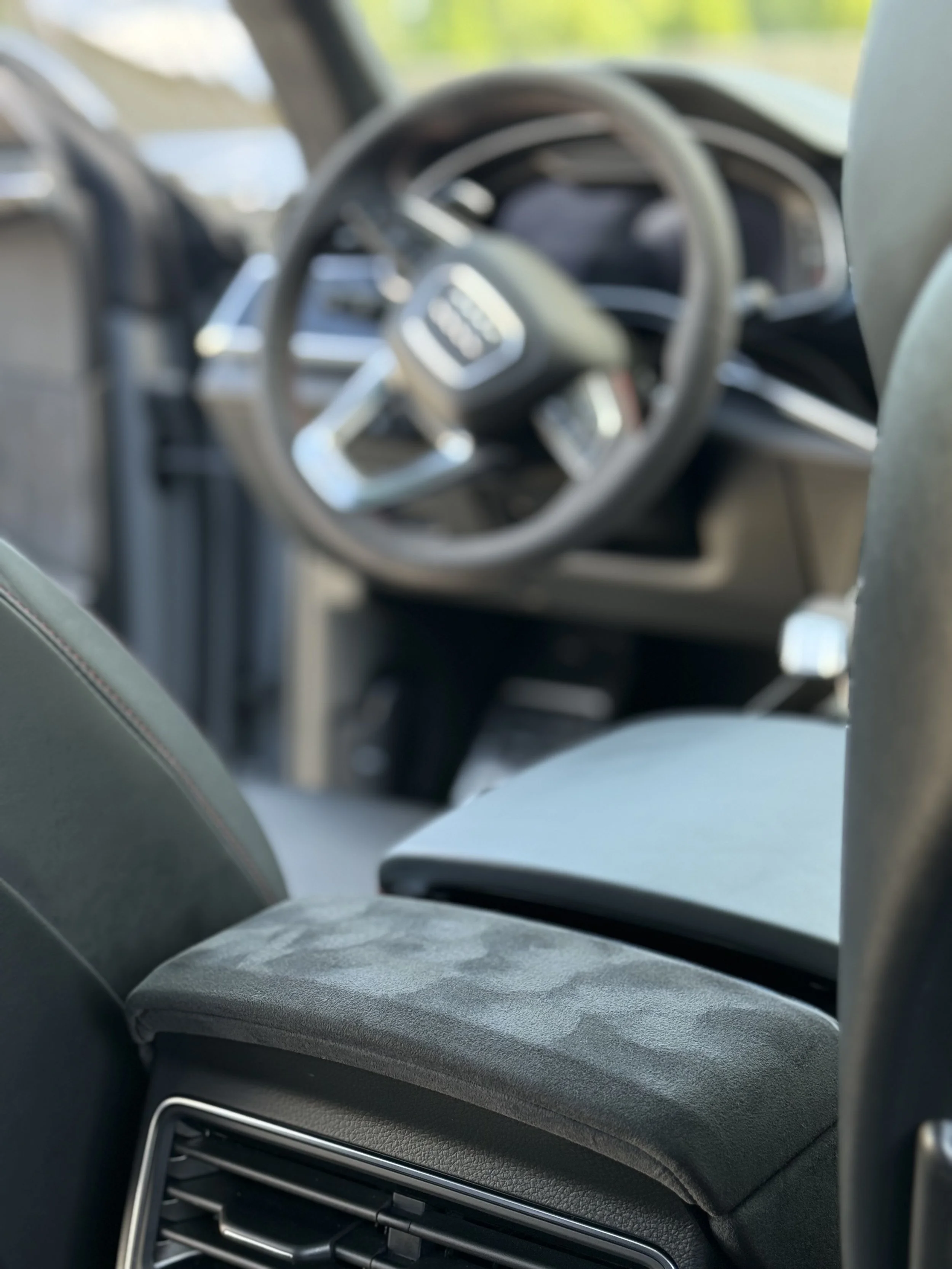 Blurred interior view of an Audi vehicle, focusing on the dashboard and steering wheel, with a gray fabric armrest in the foreground.