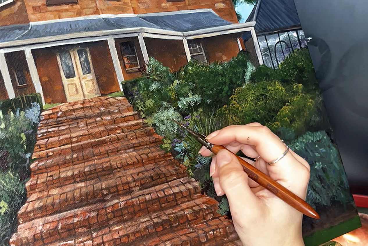 Person painting a detailed scene of a house with stairs, garden, and greenery using a fine brush.
