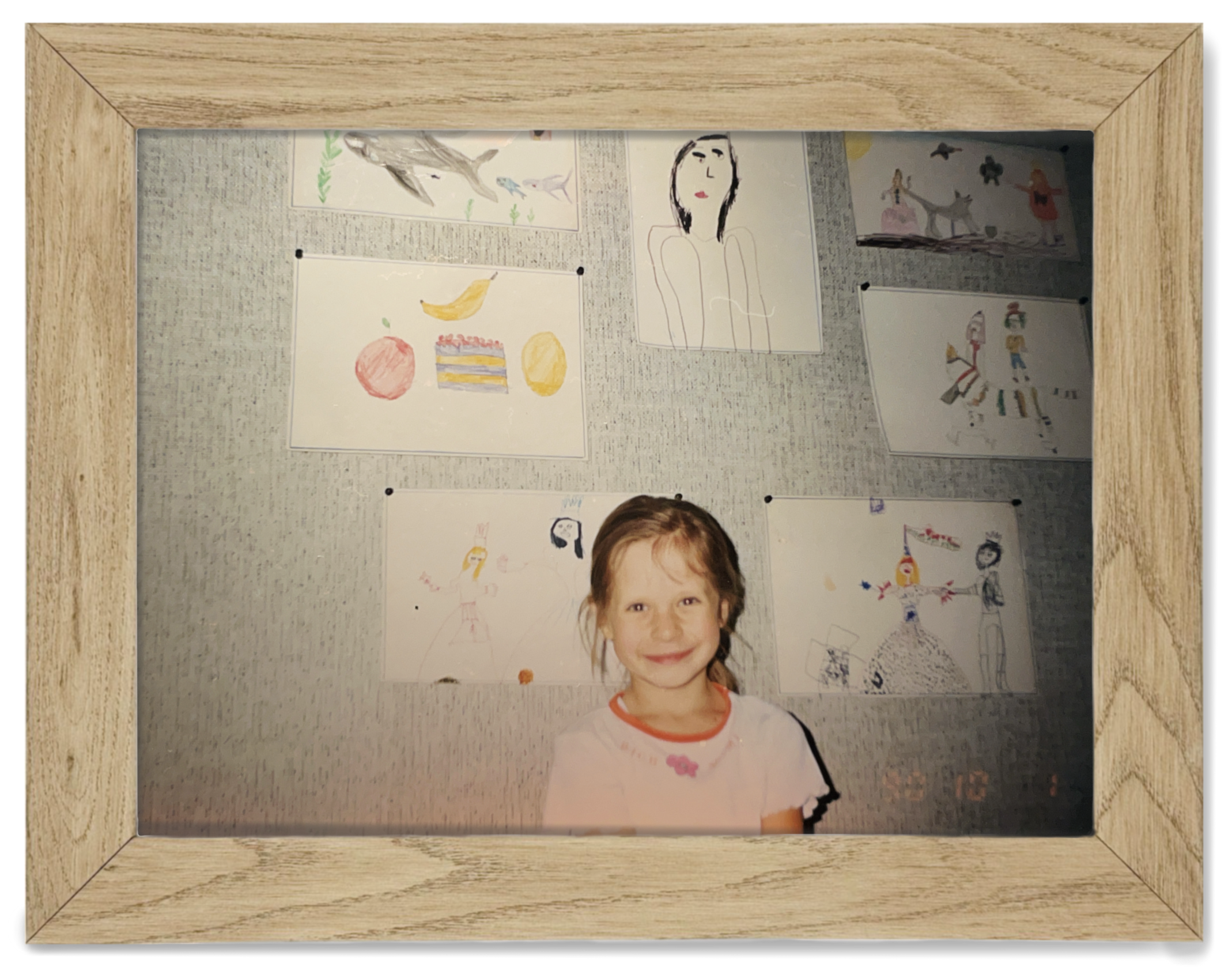 A young girl with brown hair smiling in front of a display of children's artwork on a gray wall, framed by a light wooden border.