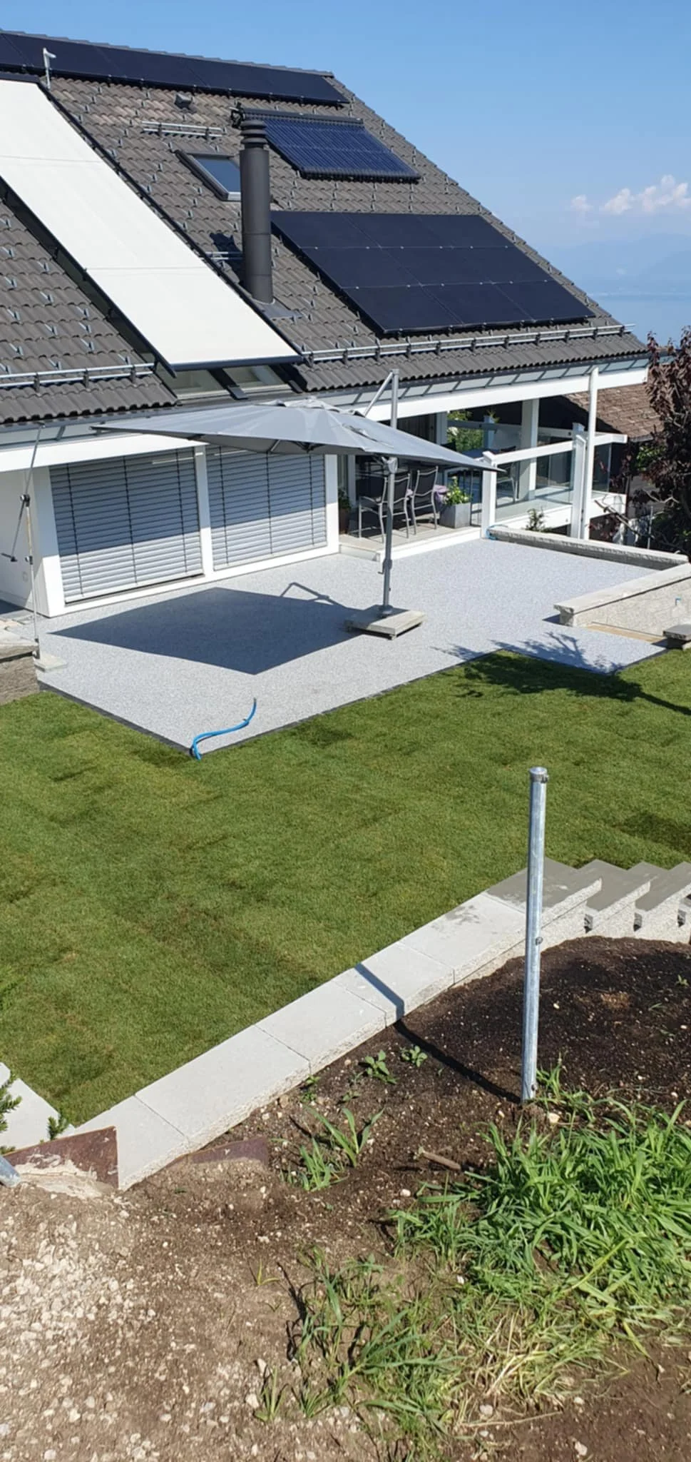 Un toit de maison avec des panneaux solaires, une terrasse avec parasol et une partie de jardin avec pelouse et parterre de fleurs.
