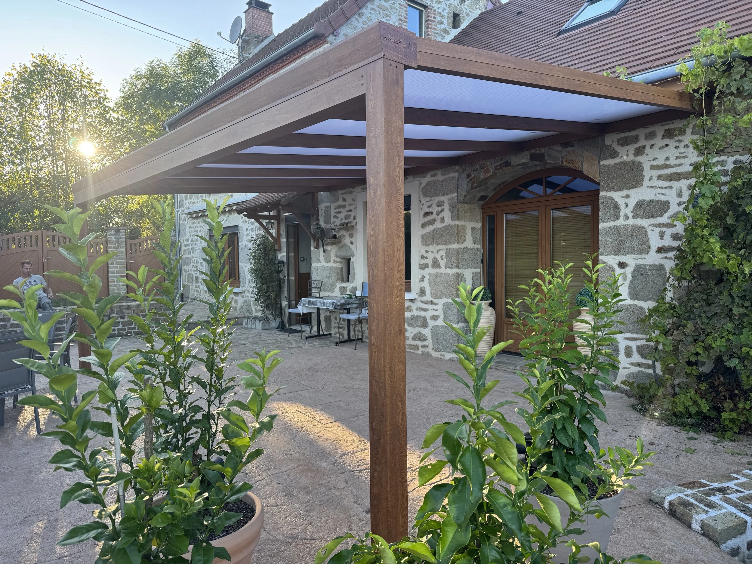 Terrasse extérieure avec pergola en bois, plantes en pot, pierres au mur, maison en pierre avec fenêtres en bois, ciel en arrière-plan.