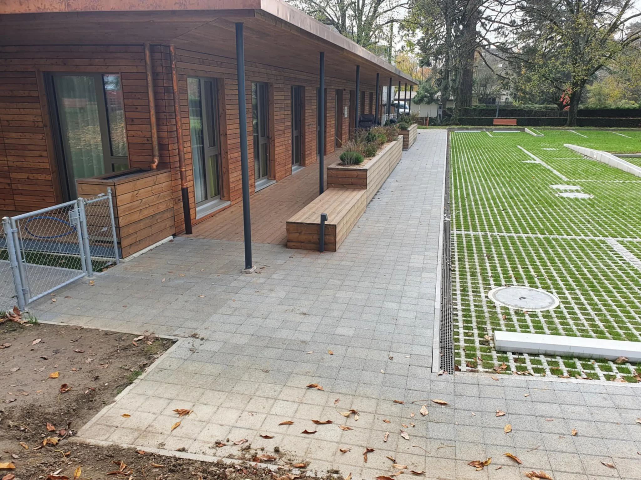 Terrasse en bois attenante à un bâtiment en bois avec bancs et jardinières, fond un terrain de sport avec pelouse synthétique, arbres et une allée de pavés.