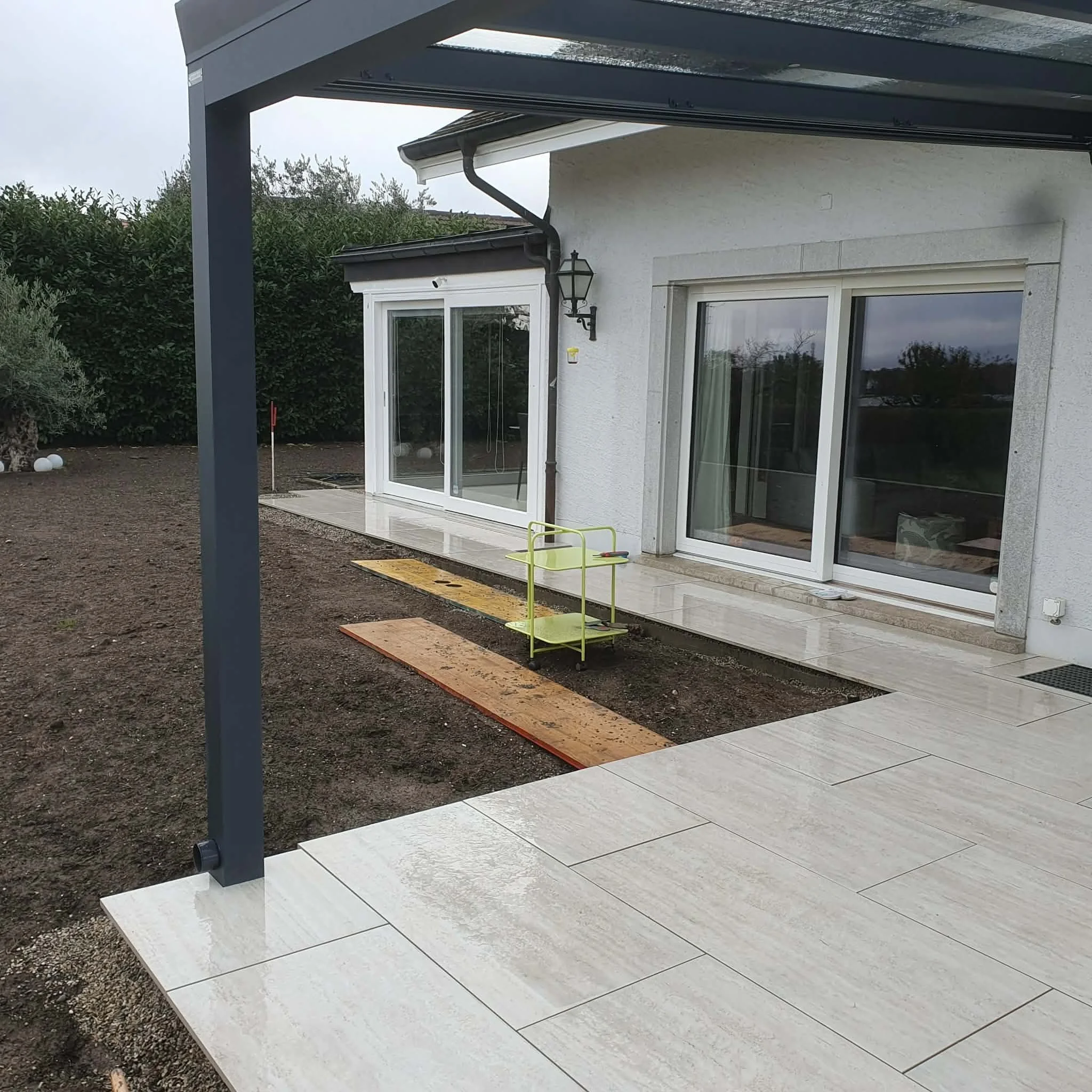 Une terrasse en pierre à l'arrière d'une maison blanche avec grandes fenêtres et porte-fenêtre, un éclairage extérieur et un ciel nuageux.