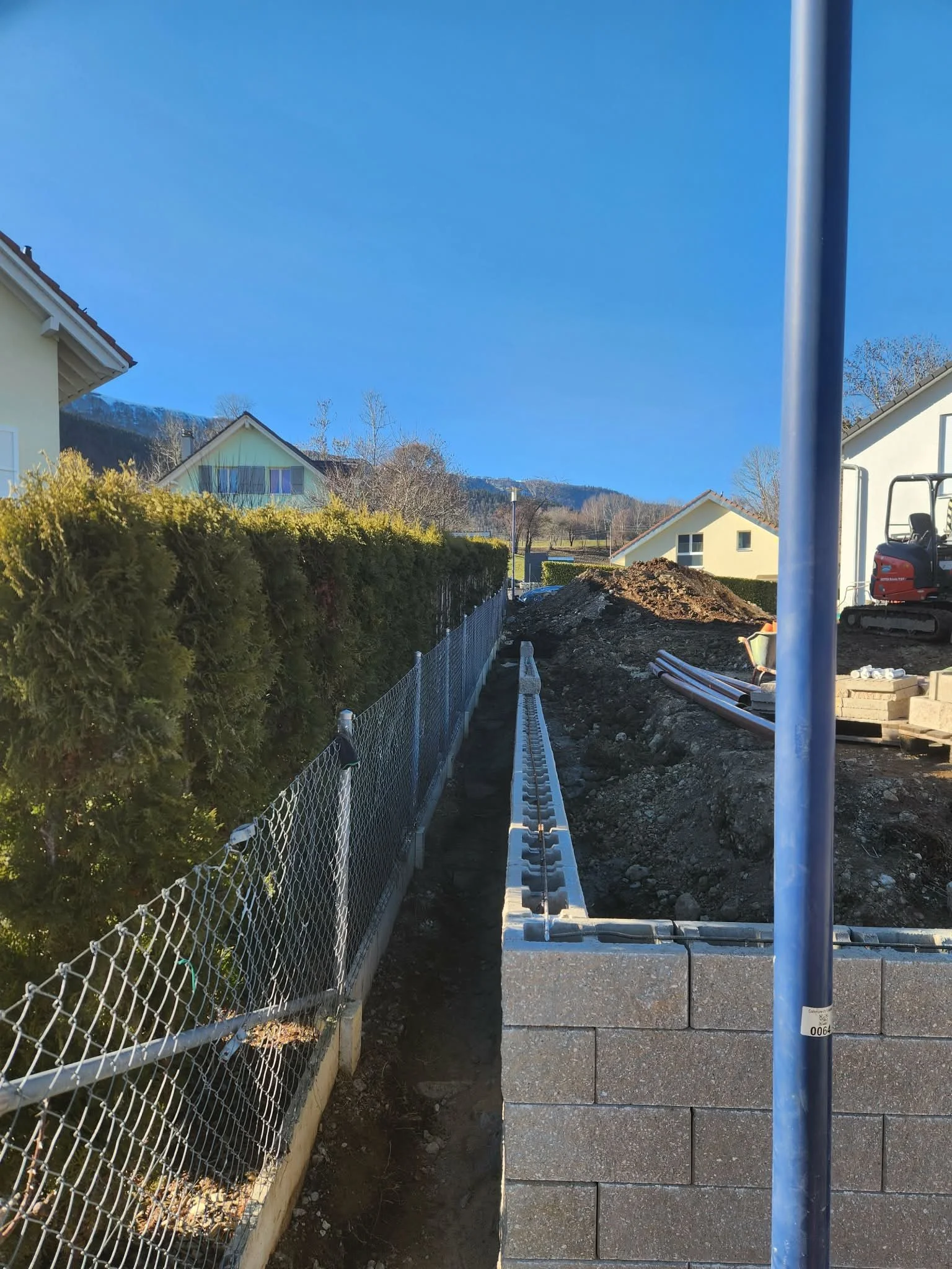 Travaux de construction avec une barrière en métal, un mur en béton, et des machines de chantier, dans une zone résidentielle.