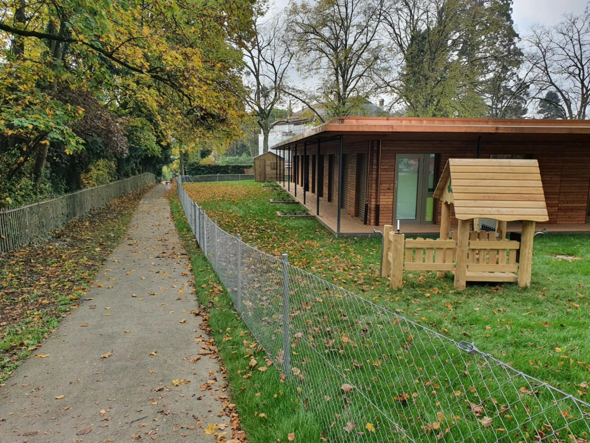 Allée bordée d'arbres avec feuilles tombées, clôture en fil métallique, bâtiment en bois avec terrasse, petit enclos en bois