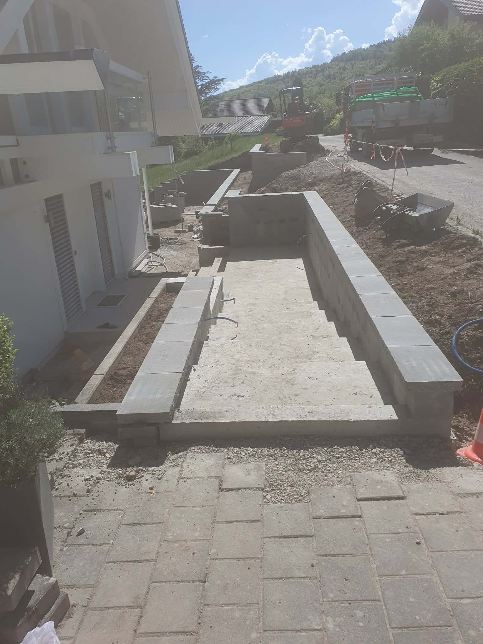 Travaux de construction d'une marche en béton avec des murs en blocs de béton à côté d'une maison. Outils et matériaux de construction visibles, avec un camion et une machine en arrière-plan, sous un ciel en partie nuageux.
