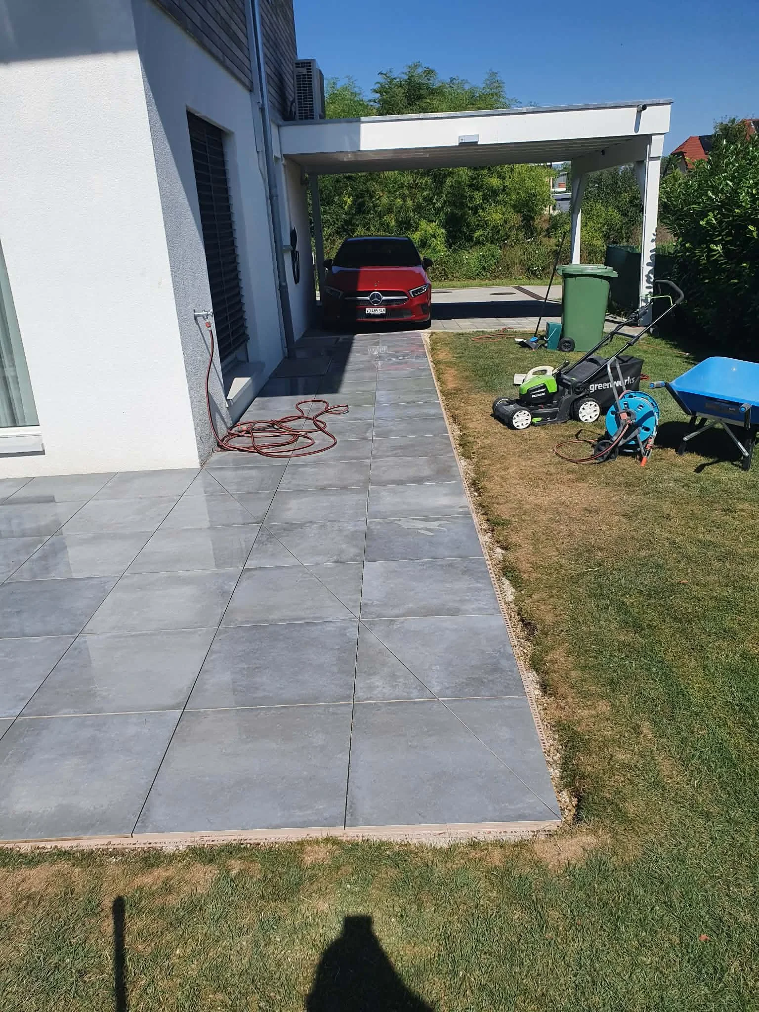 Une terrasse en carrelage devant une maison moderne, avec un garage et une voiture rouge. Sur la terrasse, des outils de jardinage et un tuyau d'arrosage. À droite, un espace gazon avec un petit camion en plastique bleu et une machine de jardinage.