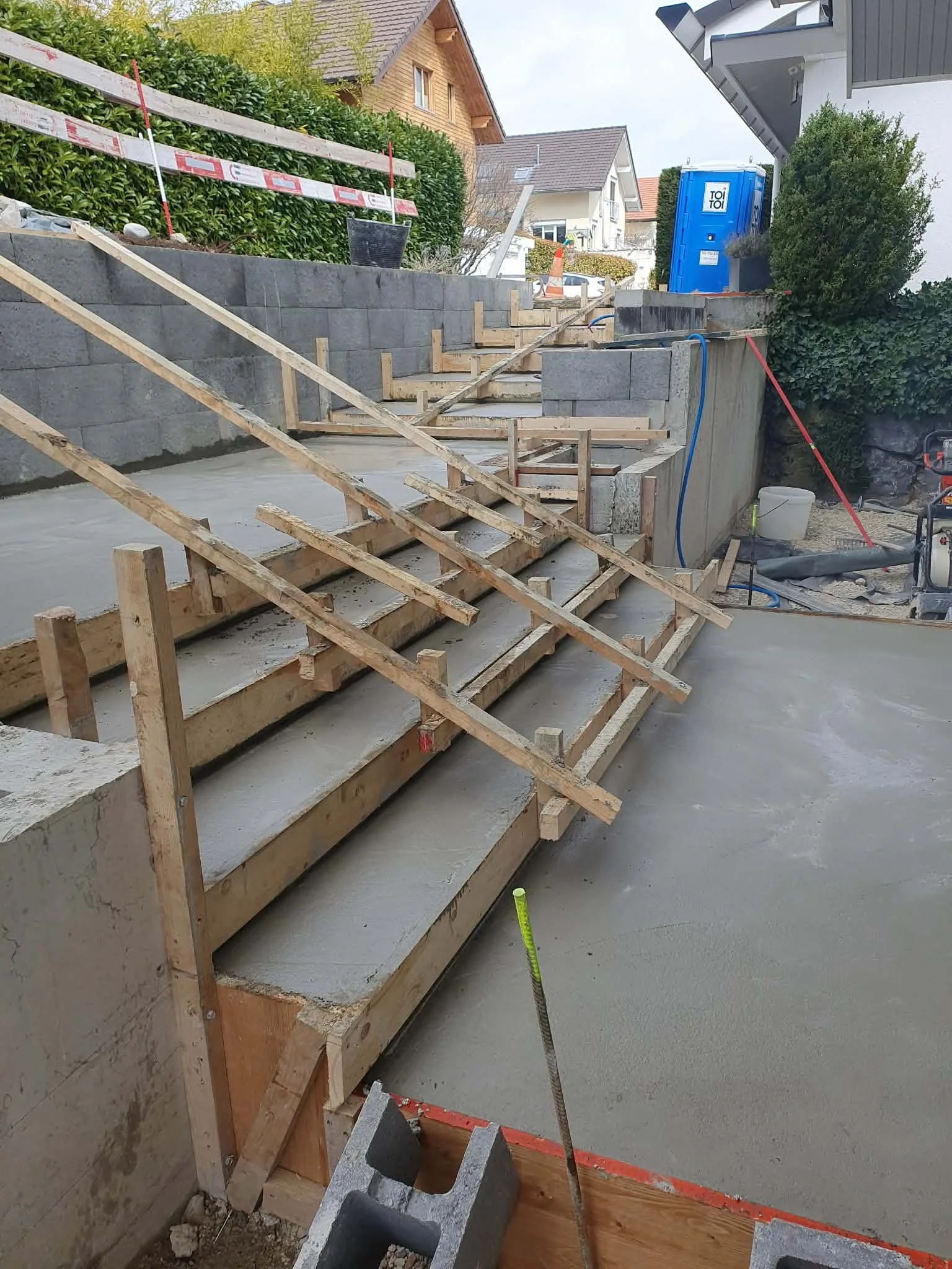 Travaux de construction d'une terrasse ou d'un escalier en béton avec échafaudages en bois et murs en pierre, dans un environnement résidentiel.
