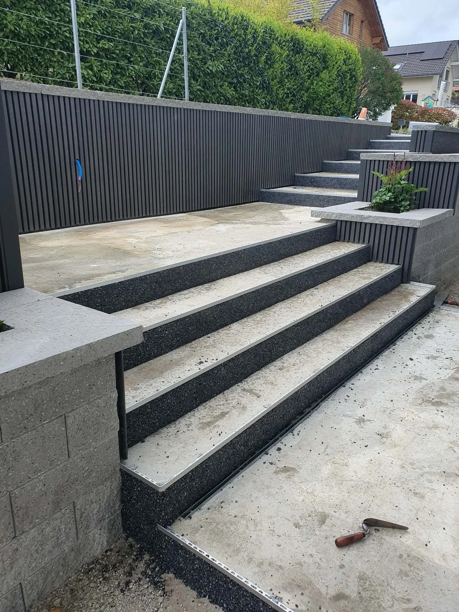 Nouveaux escaliers en construction avec des marches en pierre, entoure par des murs en panneaux noirs en aluminium, dans un paysage résidentiel.