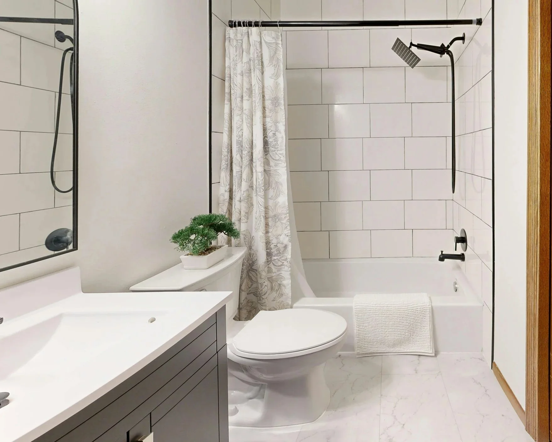 Modern bathroom with a white toilet, a vanity with a gray cabinet and white countertop, a potted green plant on the toilet tank, a bathtub with a white towel hanging over the edge, a showerhead, and a curtain rod with a white curtain in the background. The walls are light-colored with large tile accents, and the floor has a marble pattern.