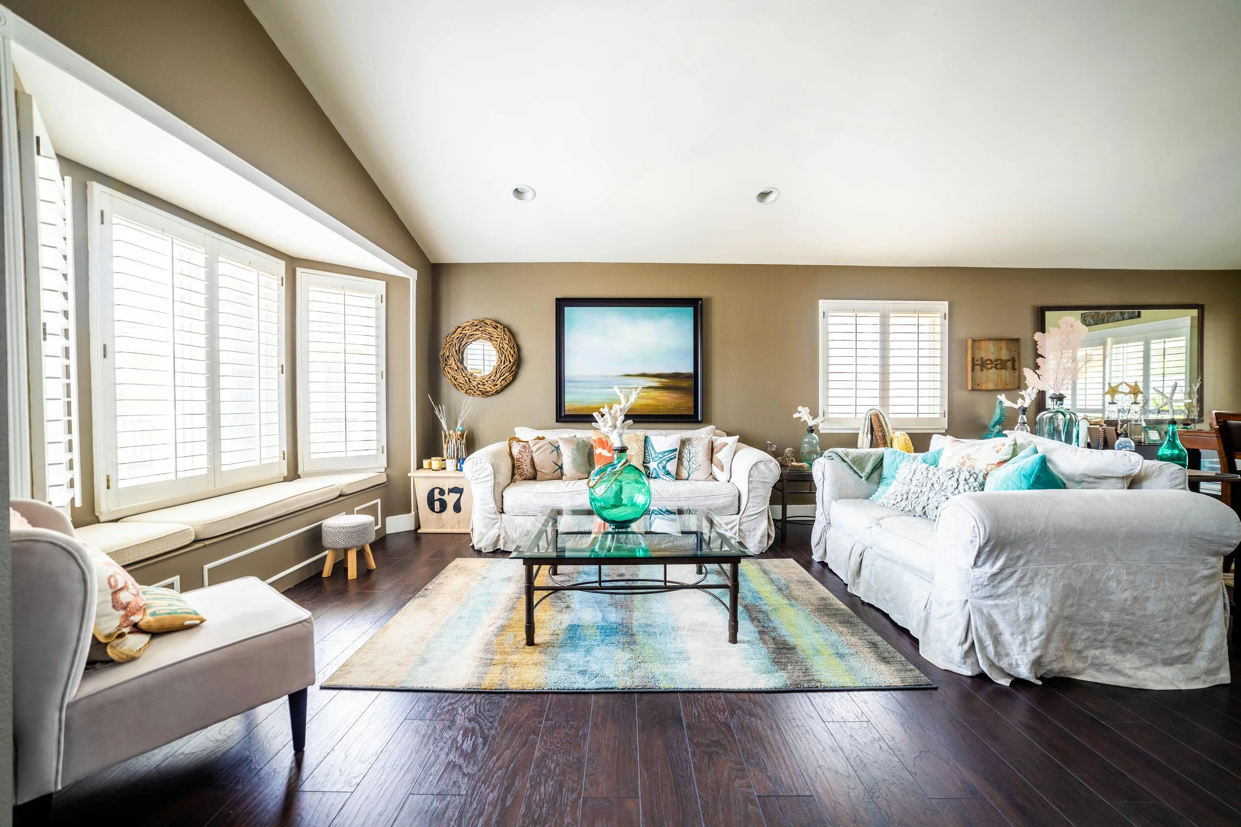 Bright living room with white couches, colorful cushions, a glass coffee table, and a nautical decor theme, featuring artwork and decorative vases, with large windows and wooden flooring.