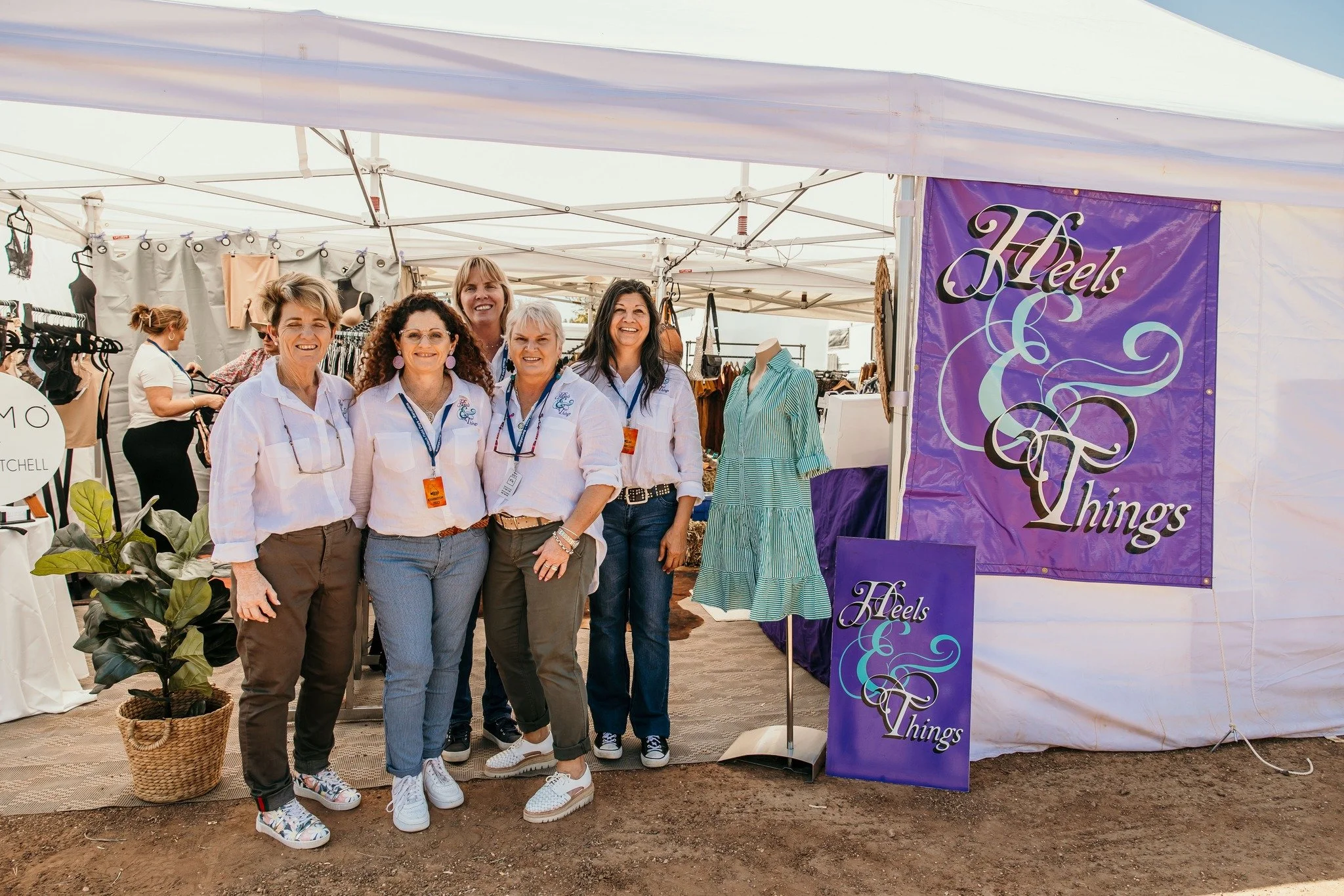 Group of six women smiling, standing in front of a booth with a purple sign that reads 'Heels & Things.' The women are wearing casual clothes and are at an outdoor event or market with other booths and mannequins in the background.