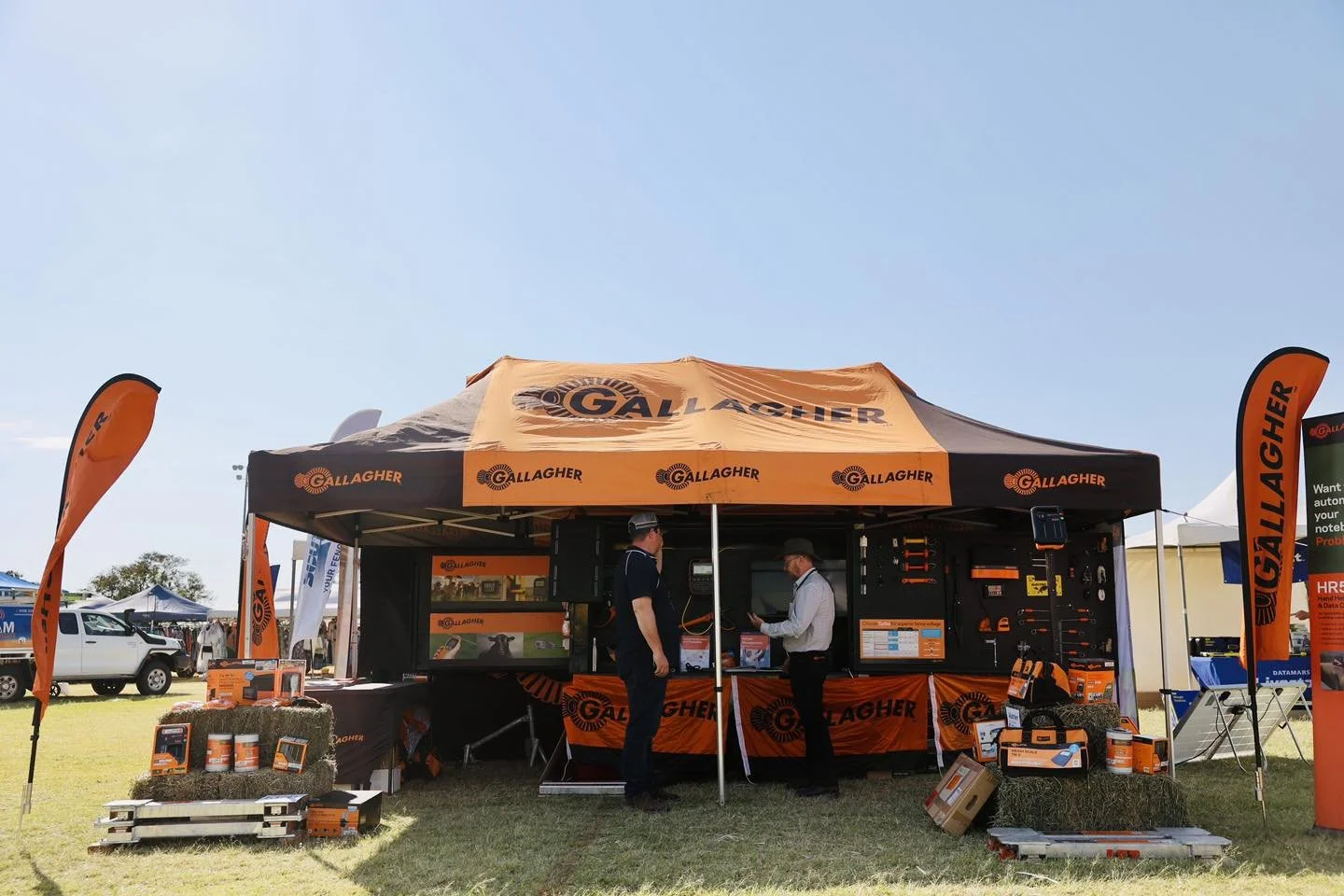 Gallagher booth at an outdoor event, with two men talking inside the tent, branded with Gallagher logos and orange and black colors, surrounded by promotional materials and equipment.