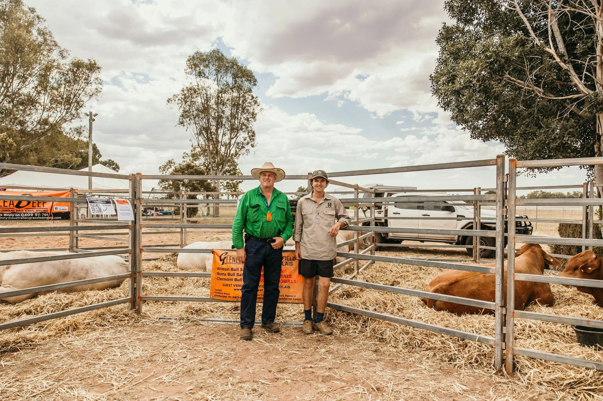 Two men standing inside a cattle pen at a livestock event. One man wears a green shirt and a cowboy hat, the other wears a beige shirt and shorts. Cattle are lying on straw inside the pen, with trees and a partly cloudy sky in the background.