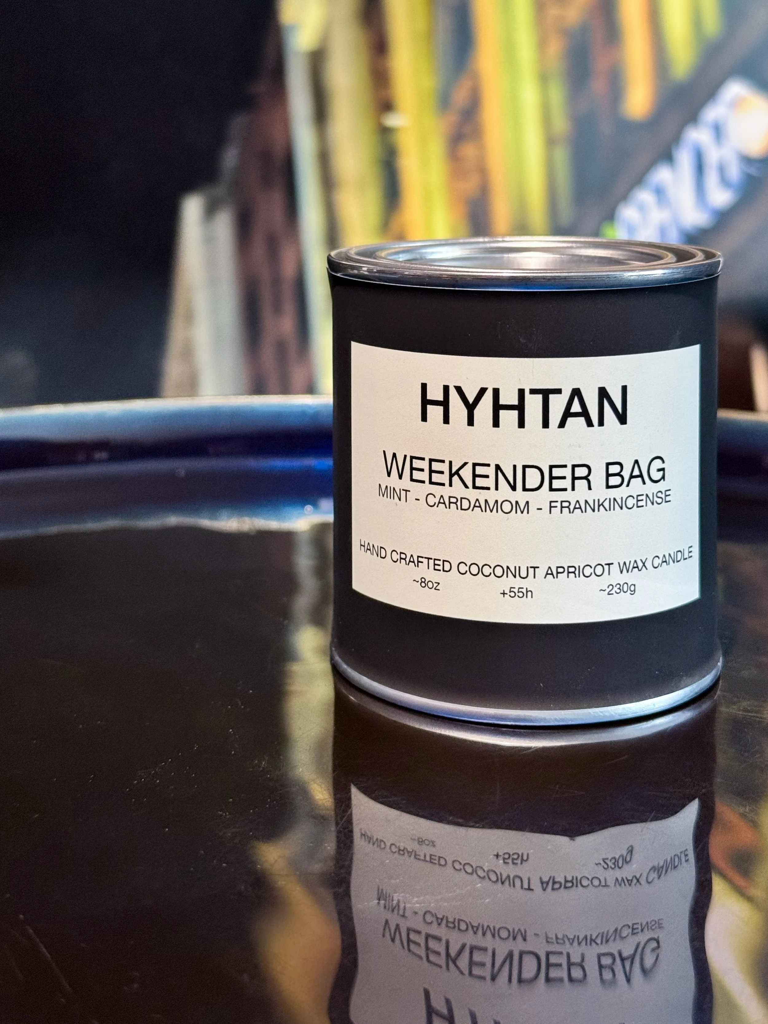 A black candle container with a white label reading 'HYHTAN WEEKENDER BAG MINT - CARDAMOM - FRANKINCENSE' sits on a reflective surface. Behind it, blurred colorful artwork is visible.