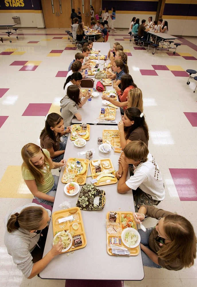 School cafeteria with children sitting at long tables, eating lunch on yellow trays with sandwiches, salads, and drinks.