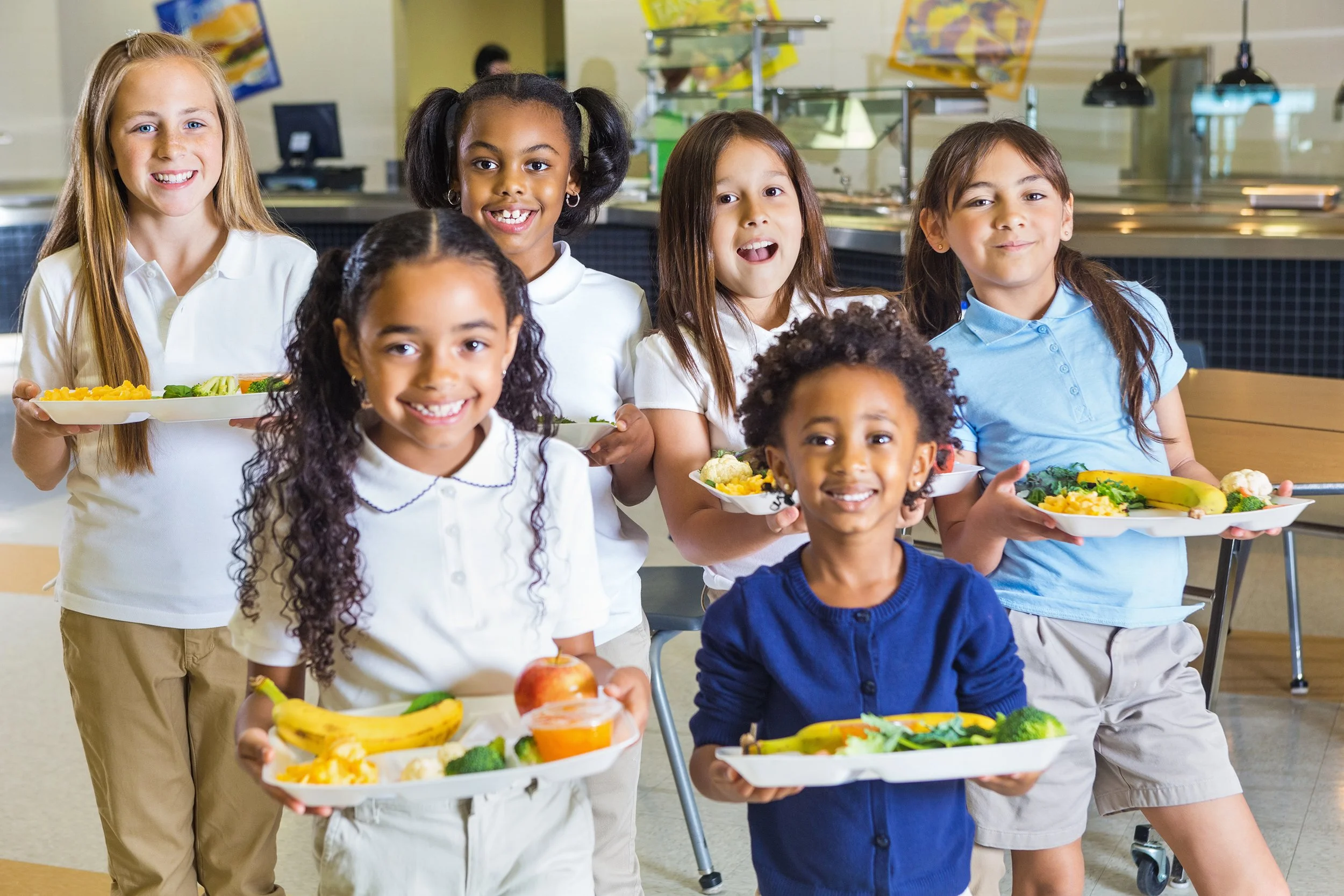 Group of six children in school uniform holding trays of food in cafeteria.