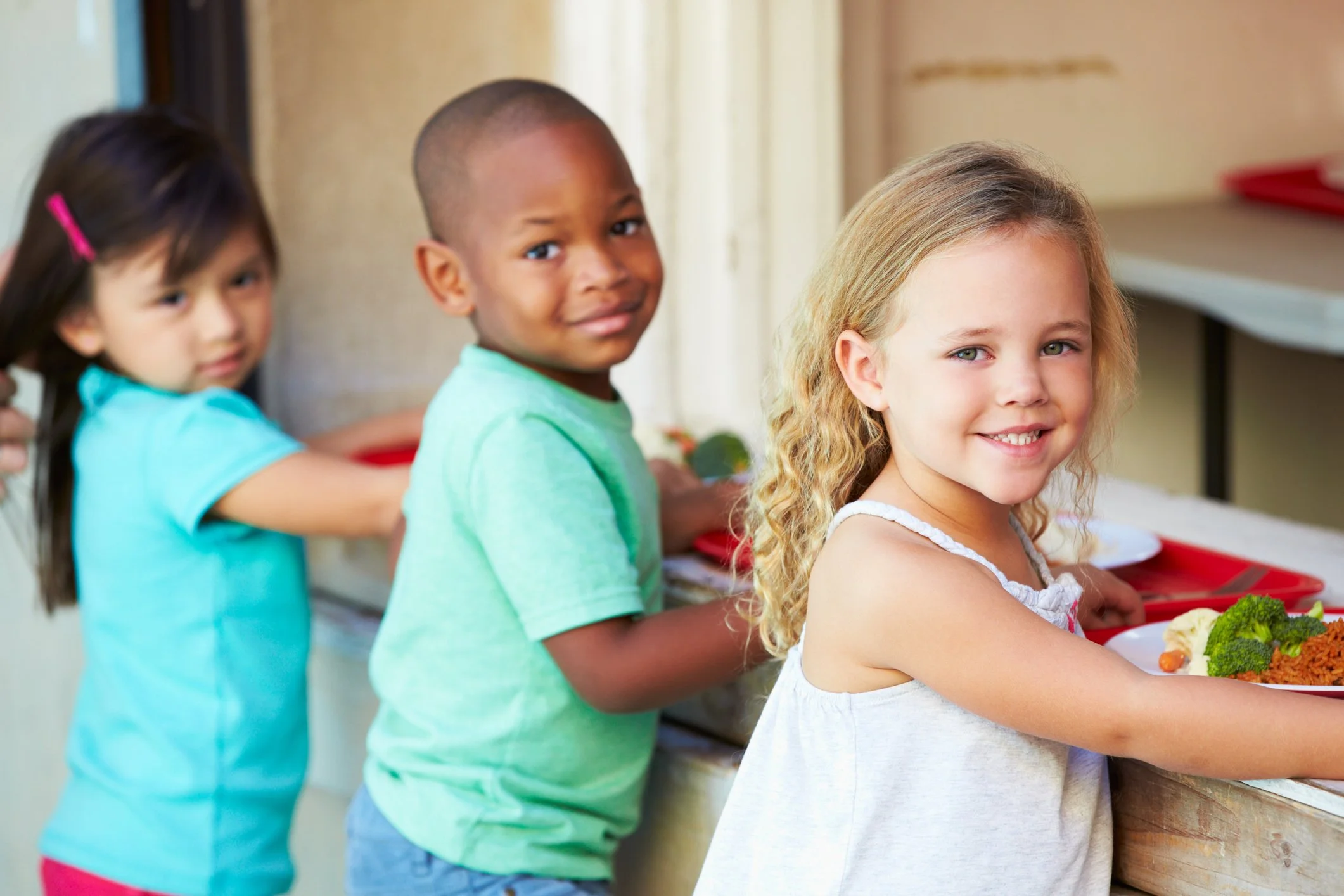 Three children standing in a line at a school lunch counter, each with a tray of food, smiling and looking at the camera.