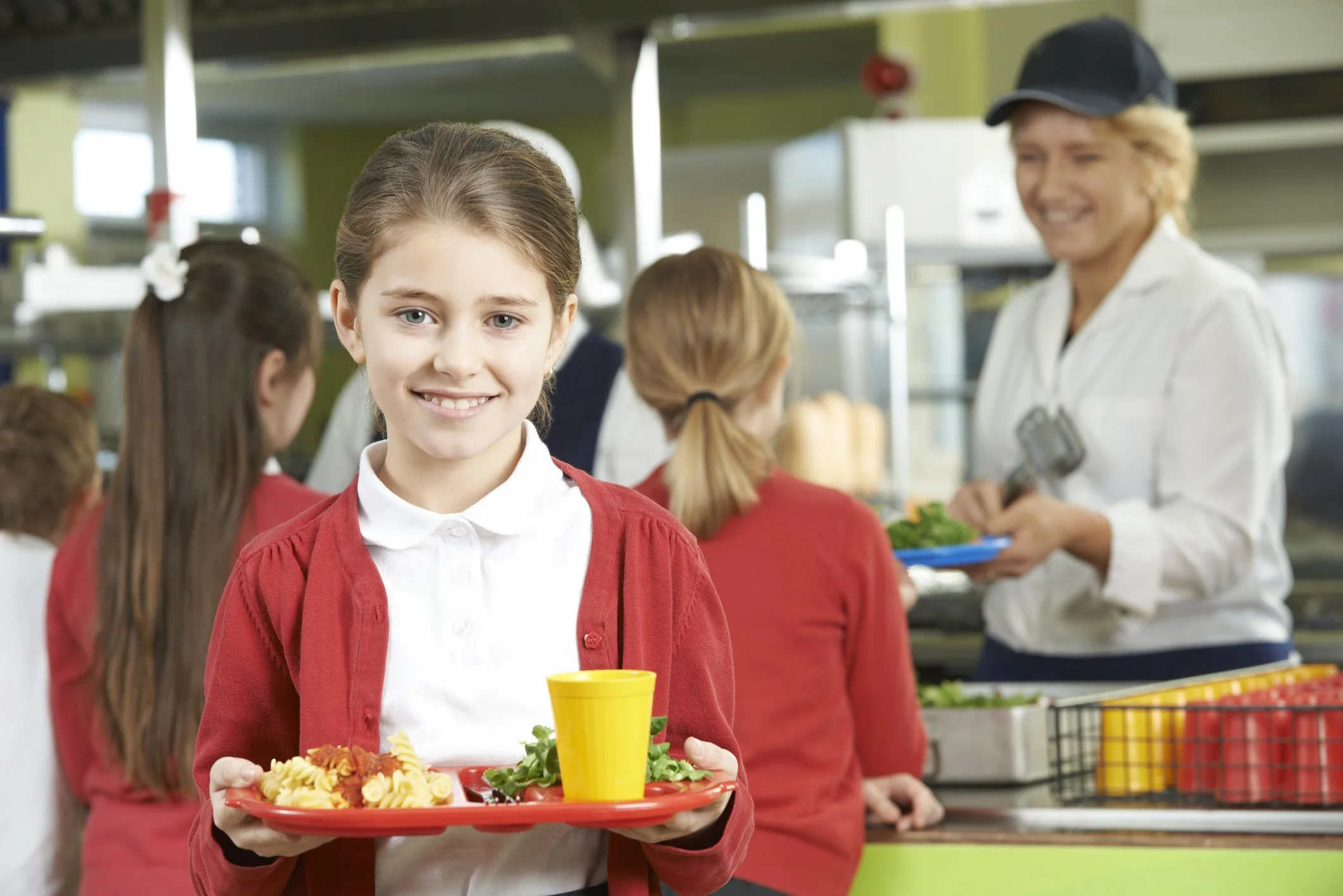 A young girl in a red school uniform holding a red tray with pasta, salad, and a yellow cup, smiling in a school cafeteria. Other children and a woman serving food are in the background.