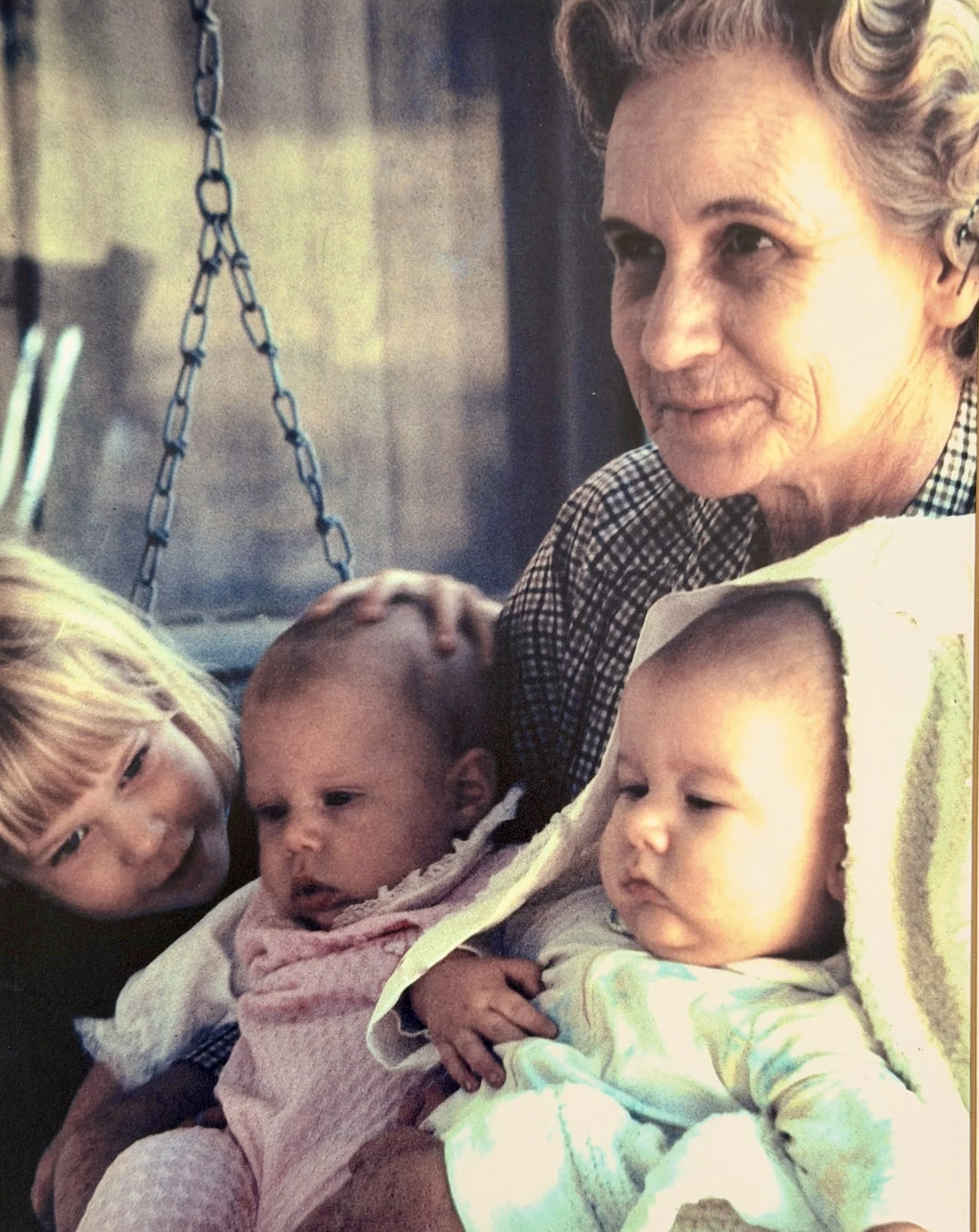 An elderly woman with gray hair sitting outside, holding three young children on her lap. One child with blonde hair and a bow, another with short hair and a pink sweater, and a third with light hair wrapped in a towel.