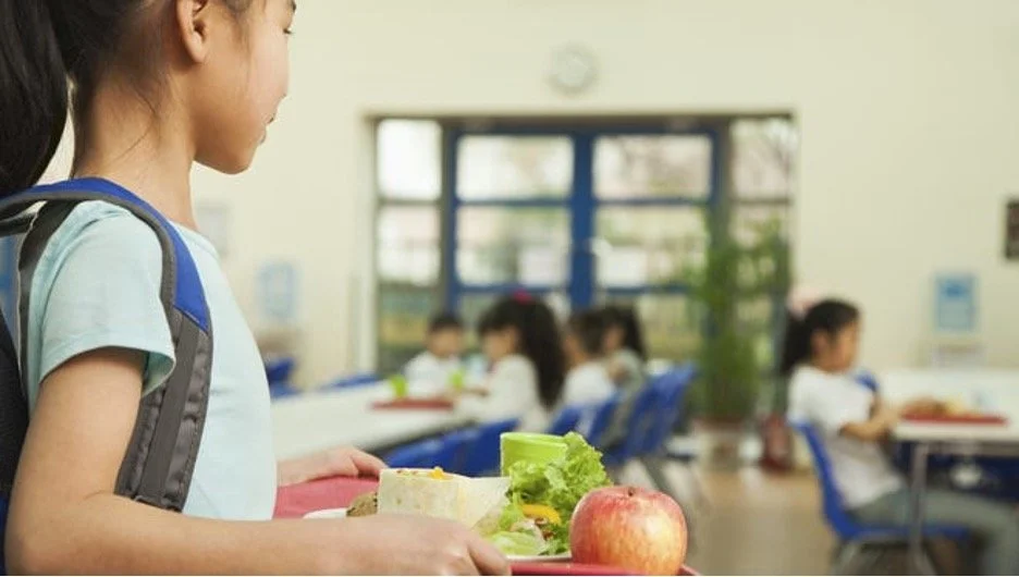 A young girl in a school cafeteria holding a tray with an apple and a sandwich, with other children sitting at tables in the background.