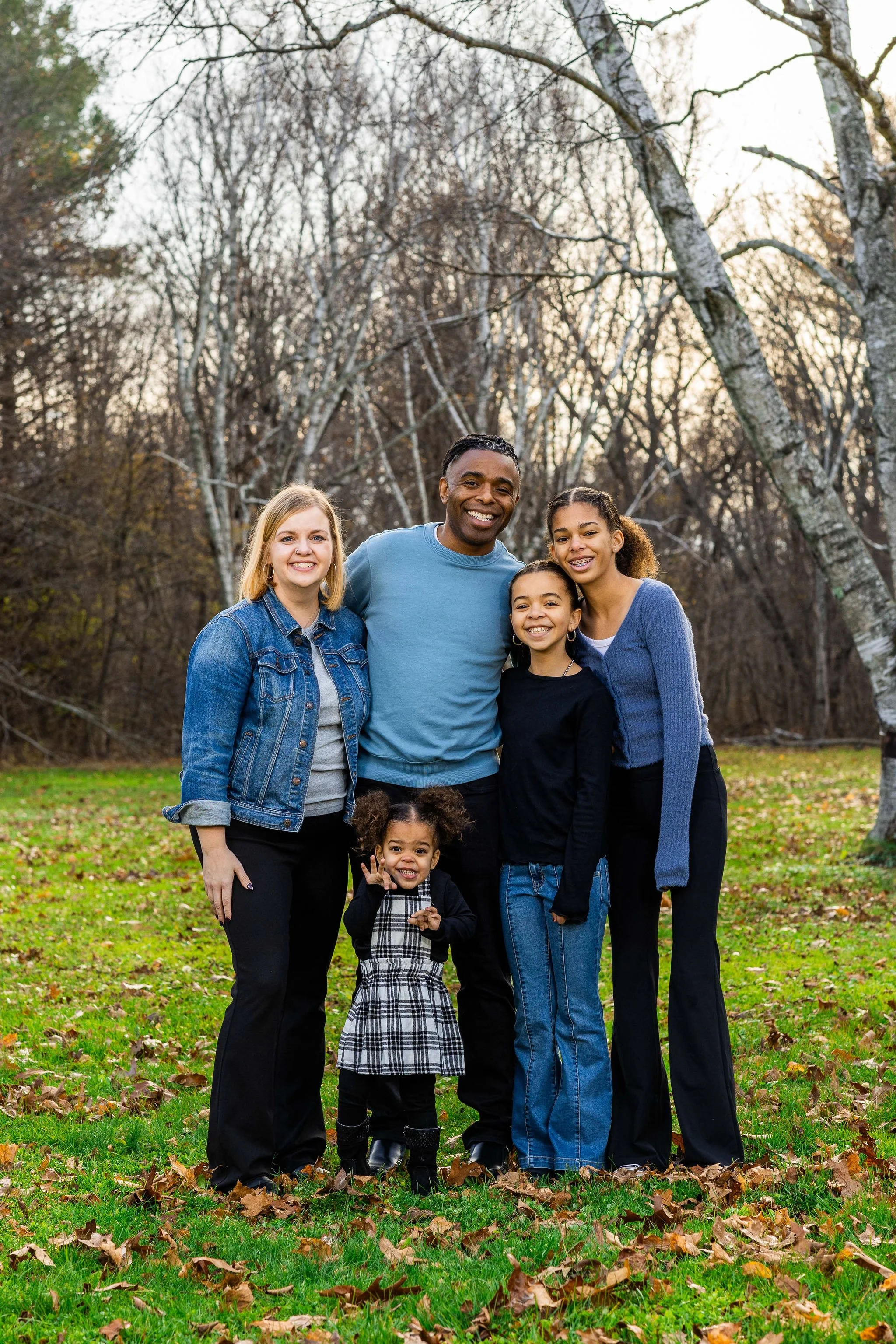 Terrence Talley and his family smiling outdoors in a park during autumn. The group includes three women, two girls, and one man, standing on grass with fallen leaves. Bare trees are in the background.