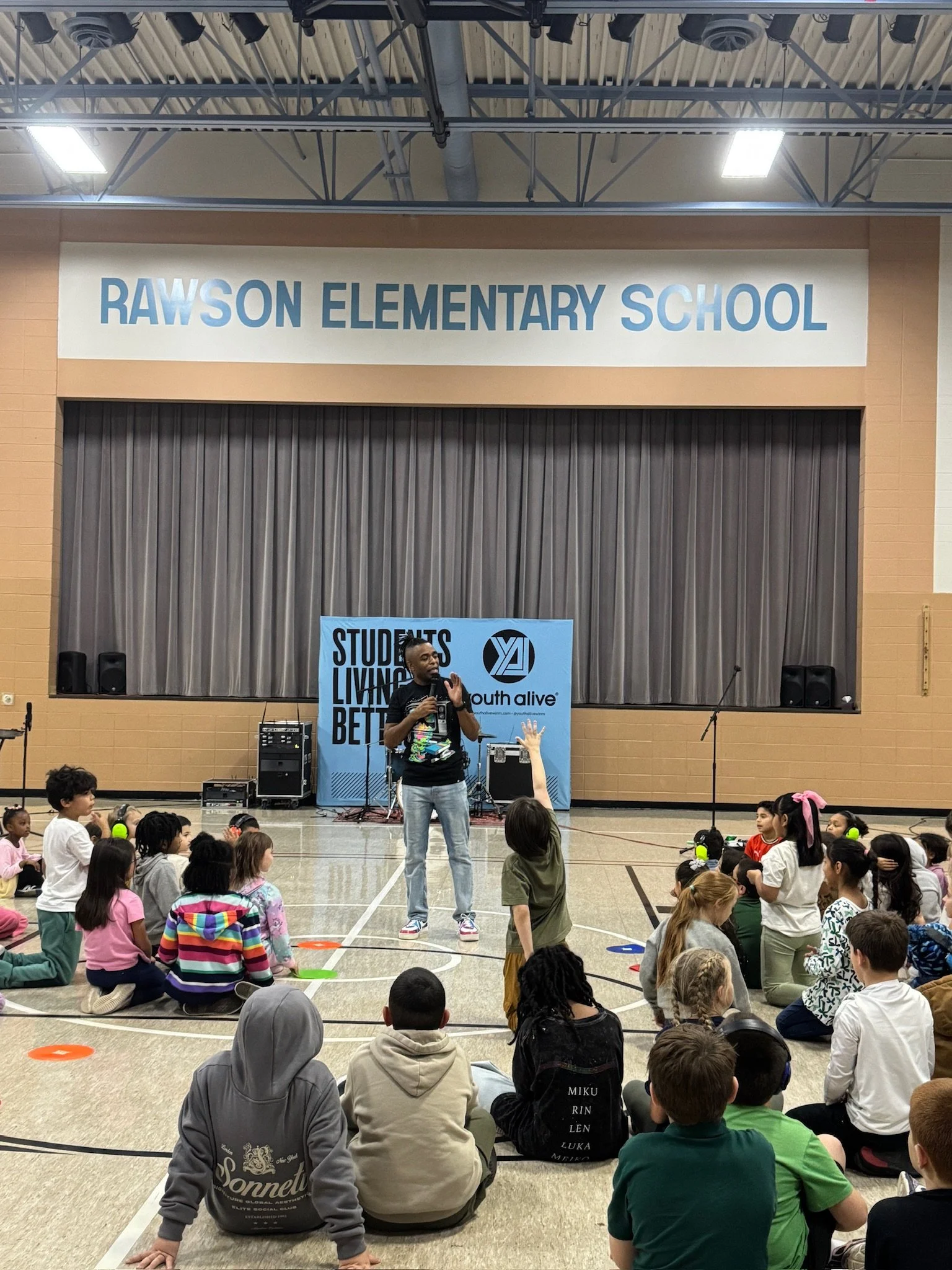Terrence Talley speaking to a group of elementary school children seated on the gym floor at Rawson Elementary School during a youth event.