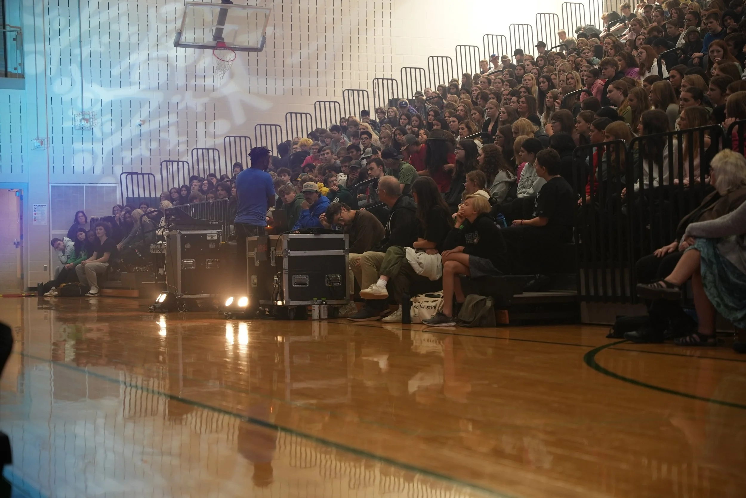 Terrence Talley in front of a School gymnasium filled with students seated on bleachers, with some sitting on the floor and a person standing near a sound or lighting equipment.
