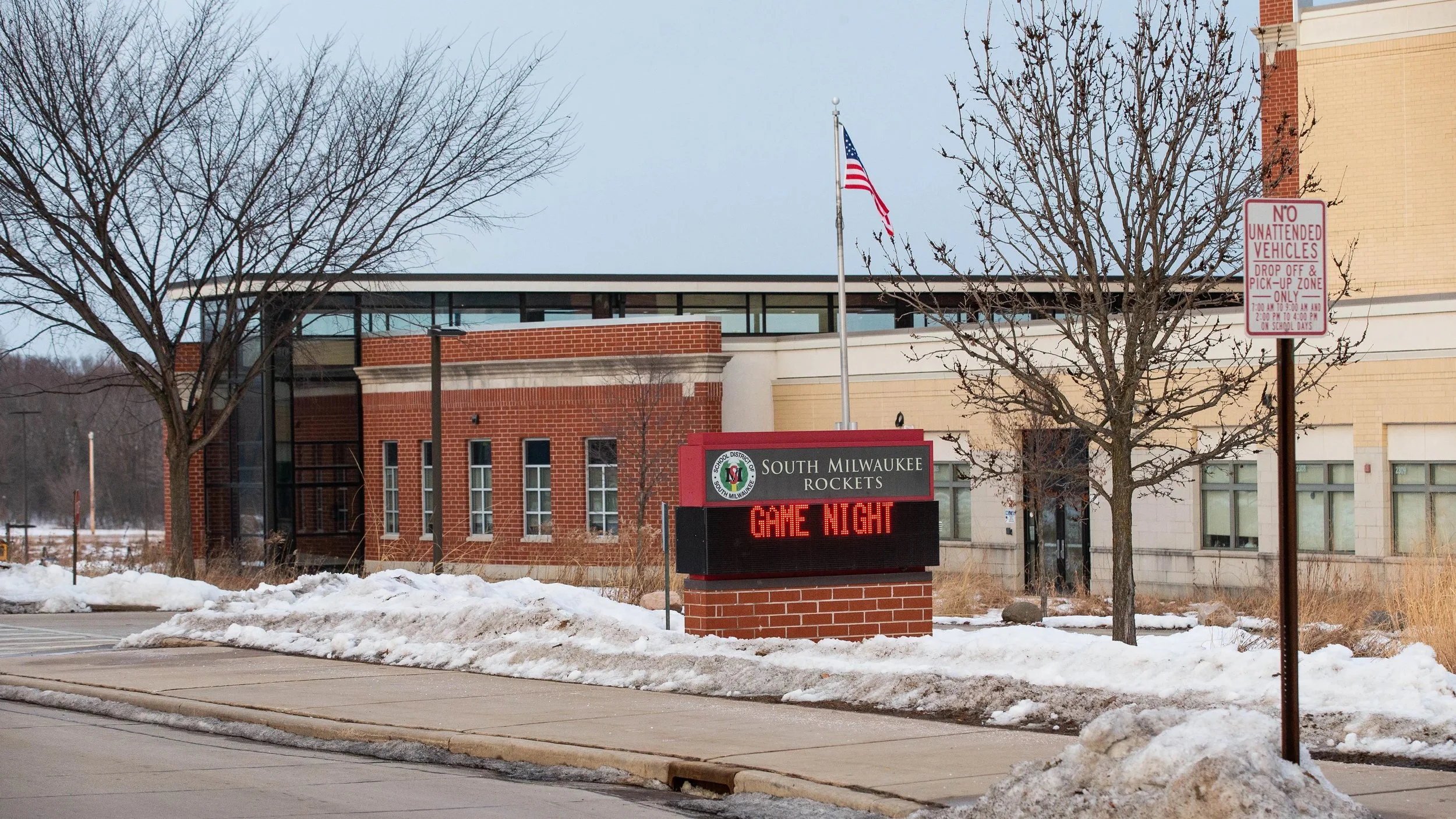 South Milwaukee School signboard with the message 'GAME NIGHT' in front of a school building, some snow on the ground, leafless trees, an American flag, and a no unauthorized vehicles sign.