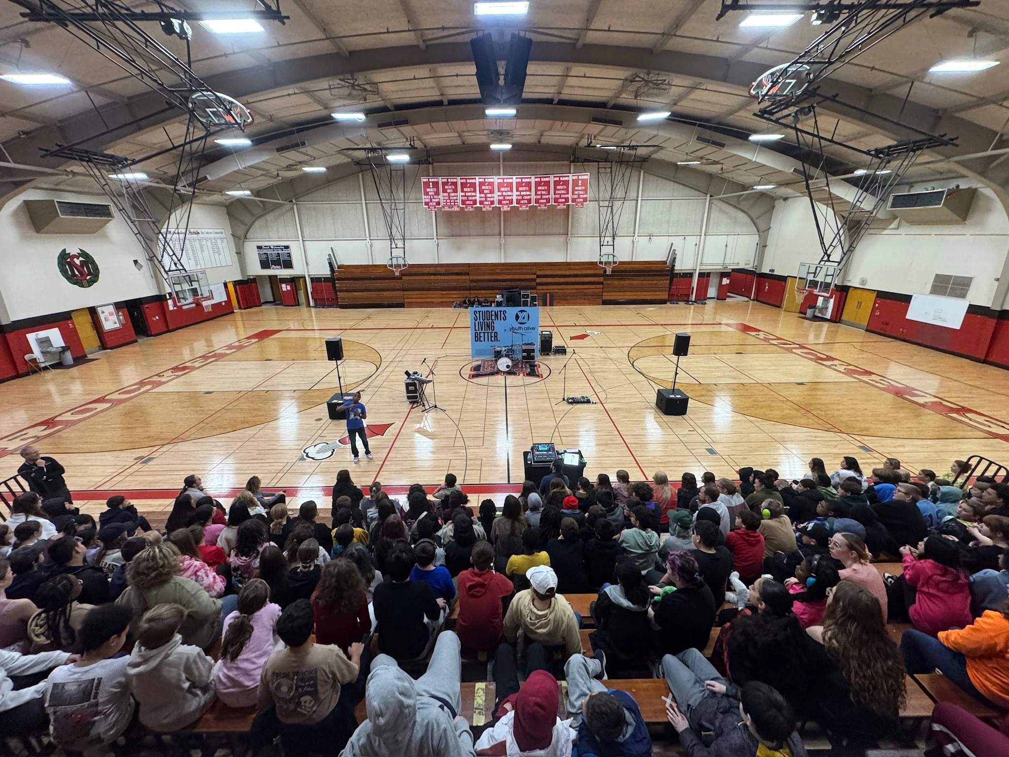 Terrence Talley in front of a group of Students attending an event in a high school gymnasium with a stage that has a backdrop reading "Students Living BETTER" and musical instruments set up at the front.