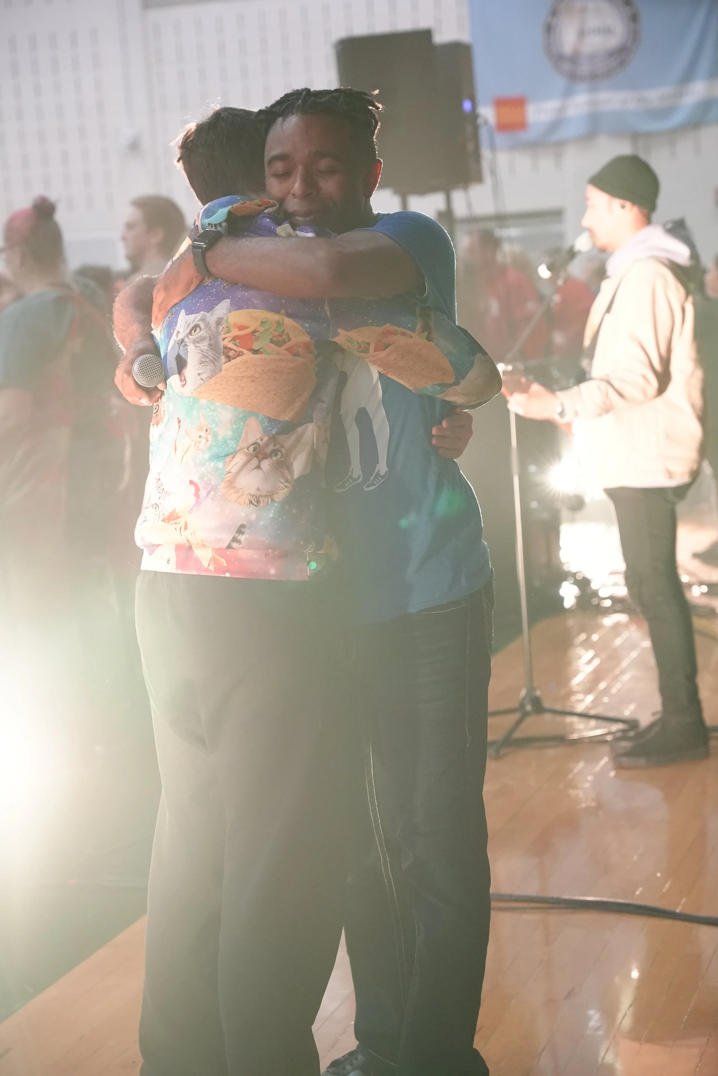Terrence Talley and a student embrace each other at a South Milwaukee indoor event. The background shows a person holding a microphone and a music stand, indicating a performance or celebration.