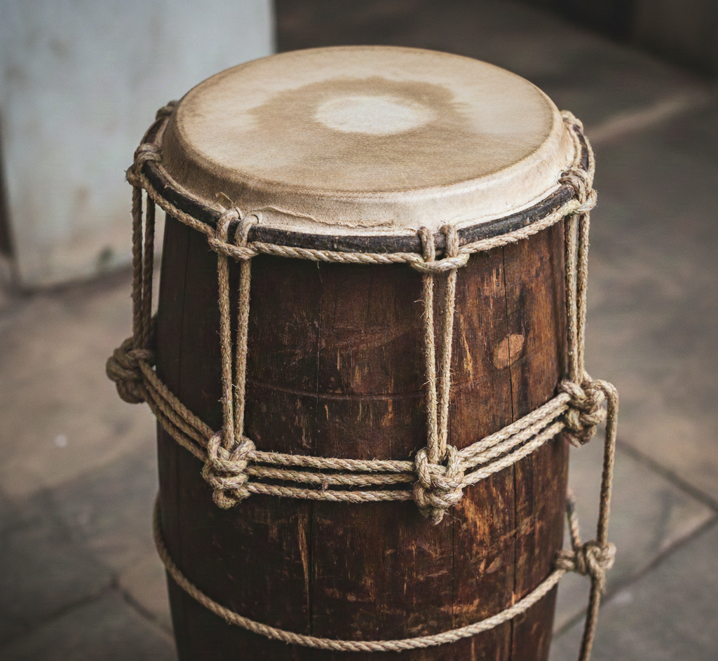 Atabaque tradicional, feito em madeira escura, com sinais de uso. Couro do instrumento com desgaste natural de uso causado pelo toque repetido nas bordas. Amarração do instrumento (afinação) feita com corda de fibra natural.