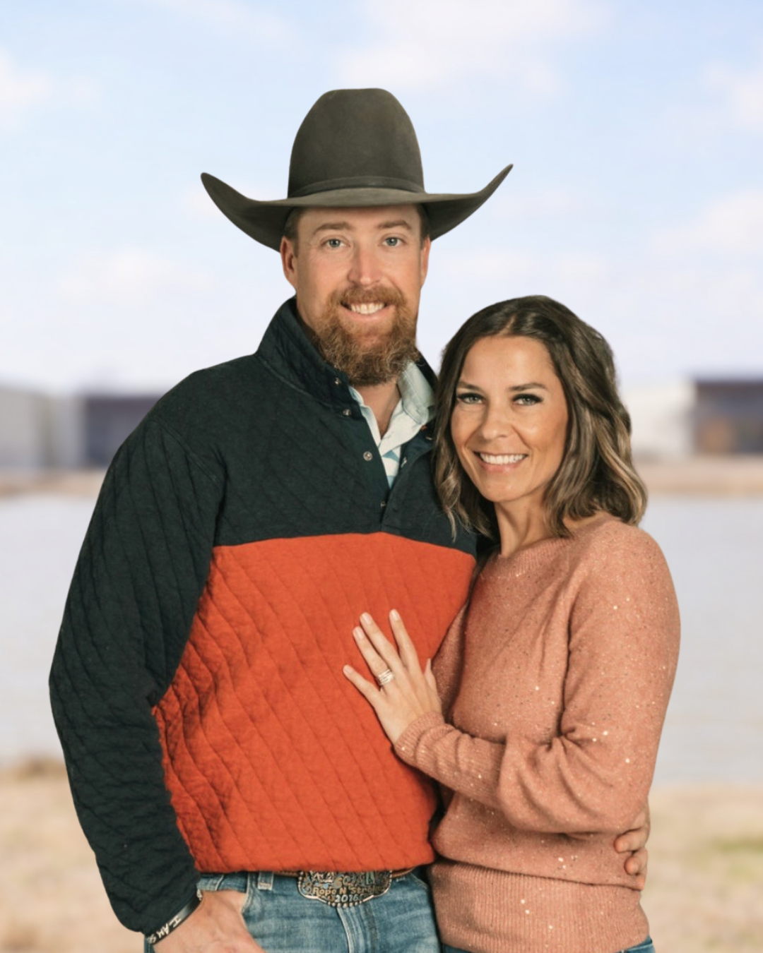 A smiling man in a cowboy hat and a woman in a pink sweater standing outdoors near water with a blurred background.