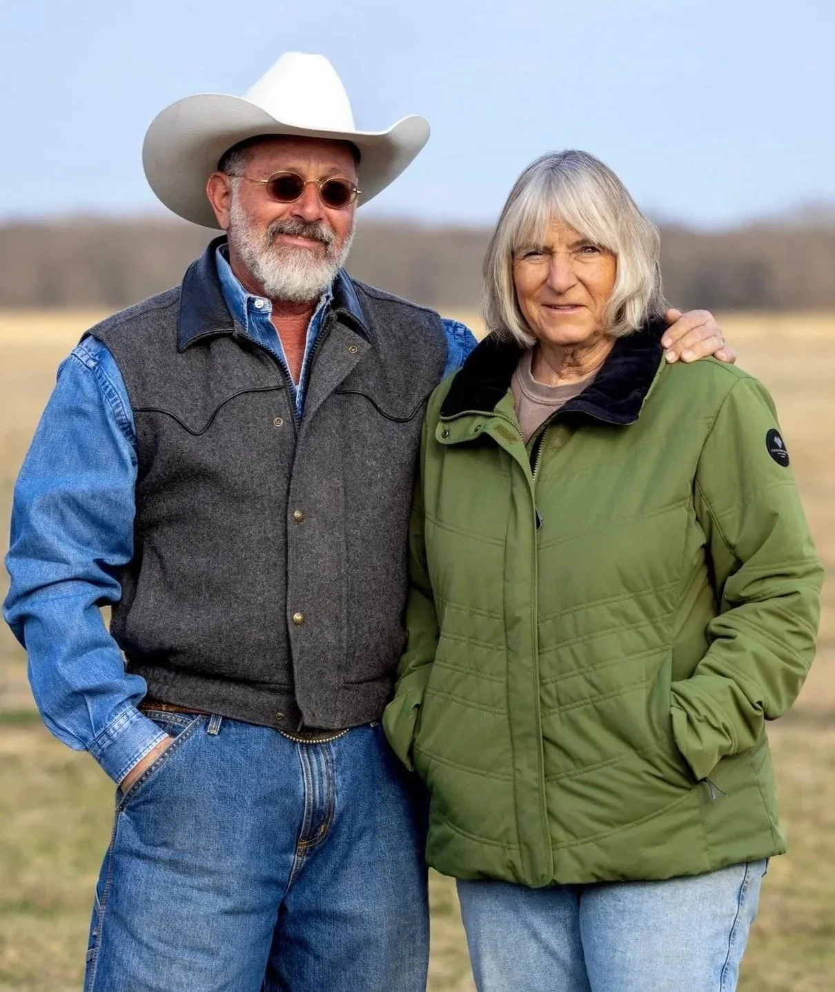 An elderly man and woman standing outdoors in a field, with the man wearing a cowboy hat, sunglasses, and a denim and fleece vest, while the woman wears a green jacket. The man has his arm around the woman.