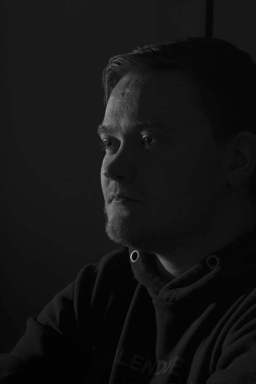 Black and white photo of a young man with short hair, looking to the side, with shadows covering most of his face, wearing a hoodie.