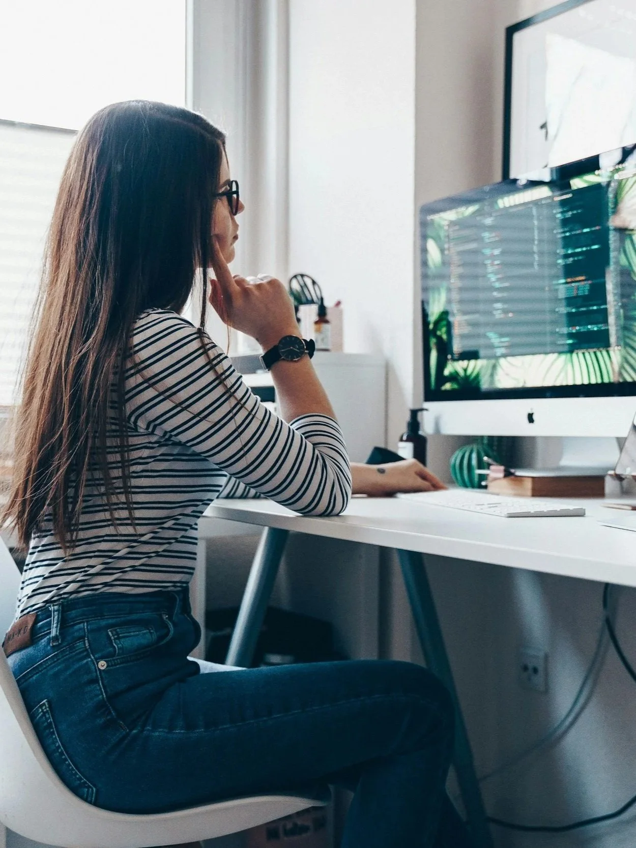 Woman with long brown hair, wearing glasses, a striped shirt, and jeans, sitting at a white desk working on a computer with a large monitor, in a well-lit room.