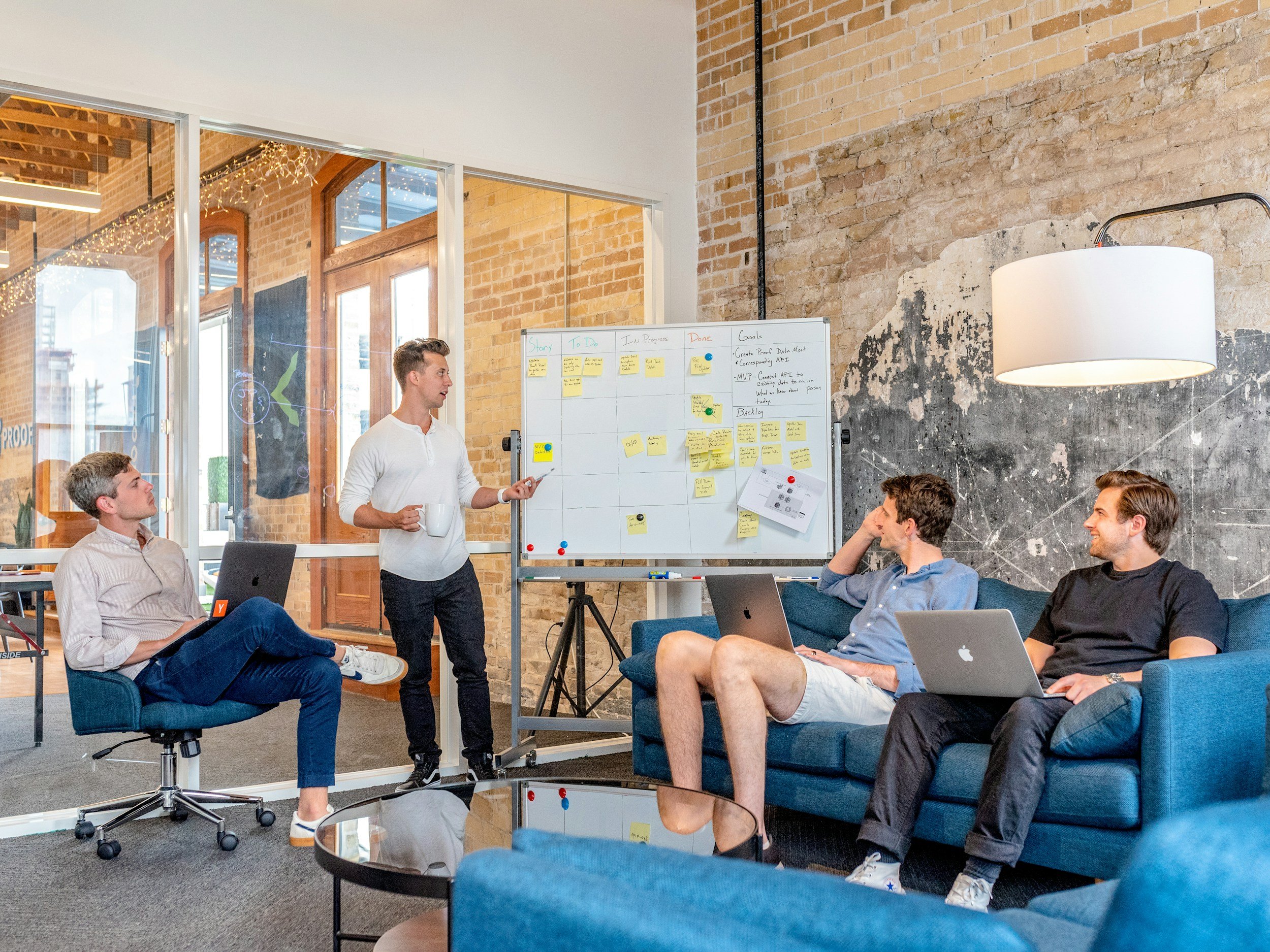 Four young men having a meeting in an office with exposed brick walls, a dry-erase board with sticky notes, and laptops.