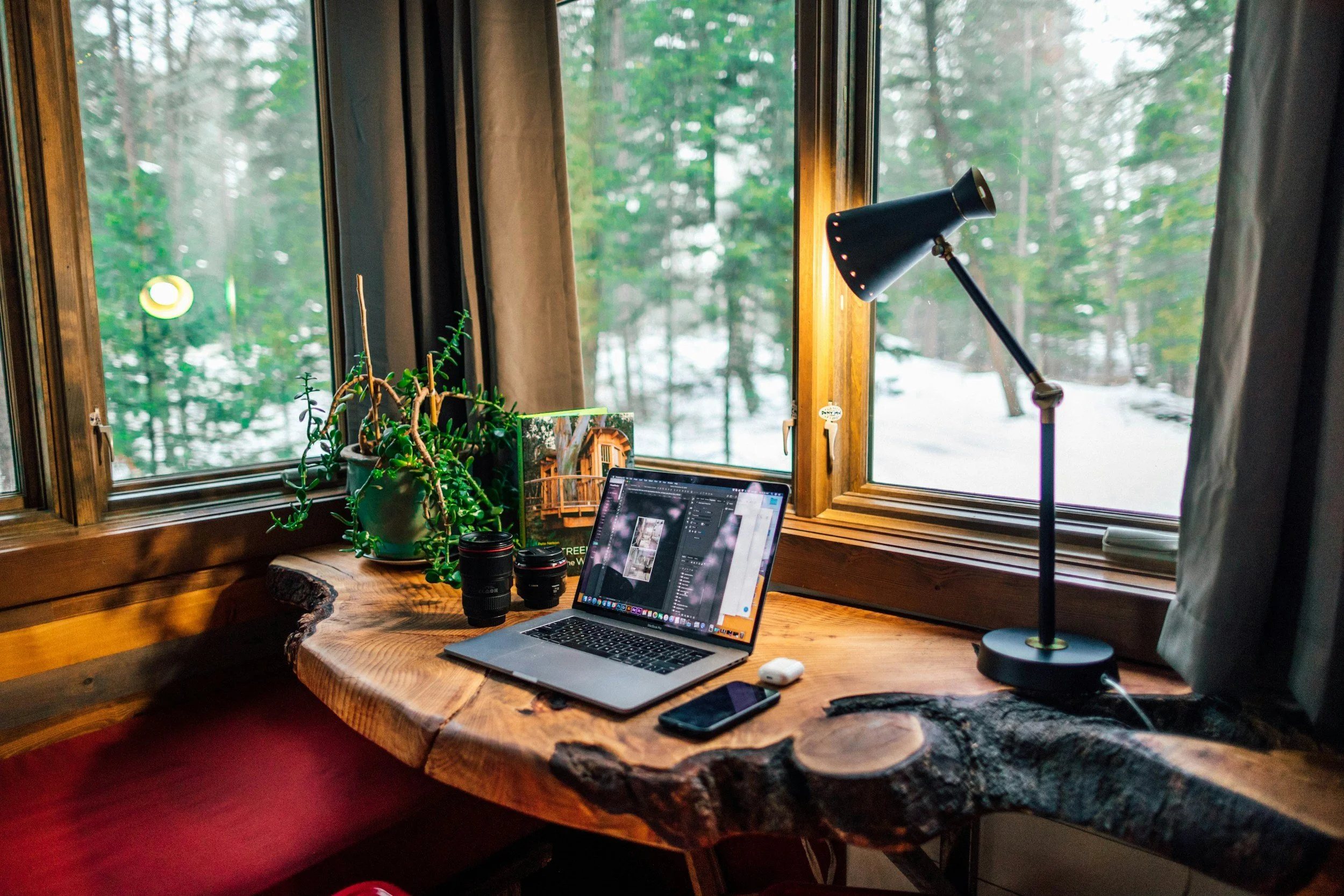Wooden desk with a laptop, smartphone, camera lenses, and a table lamp, placed in front of windows showing a snowy outdoor forest.