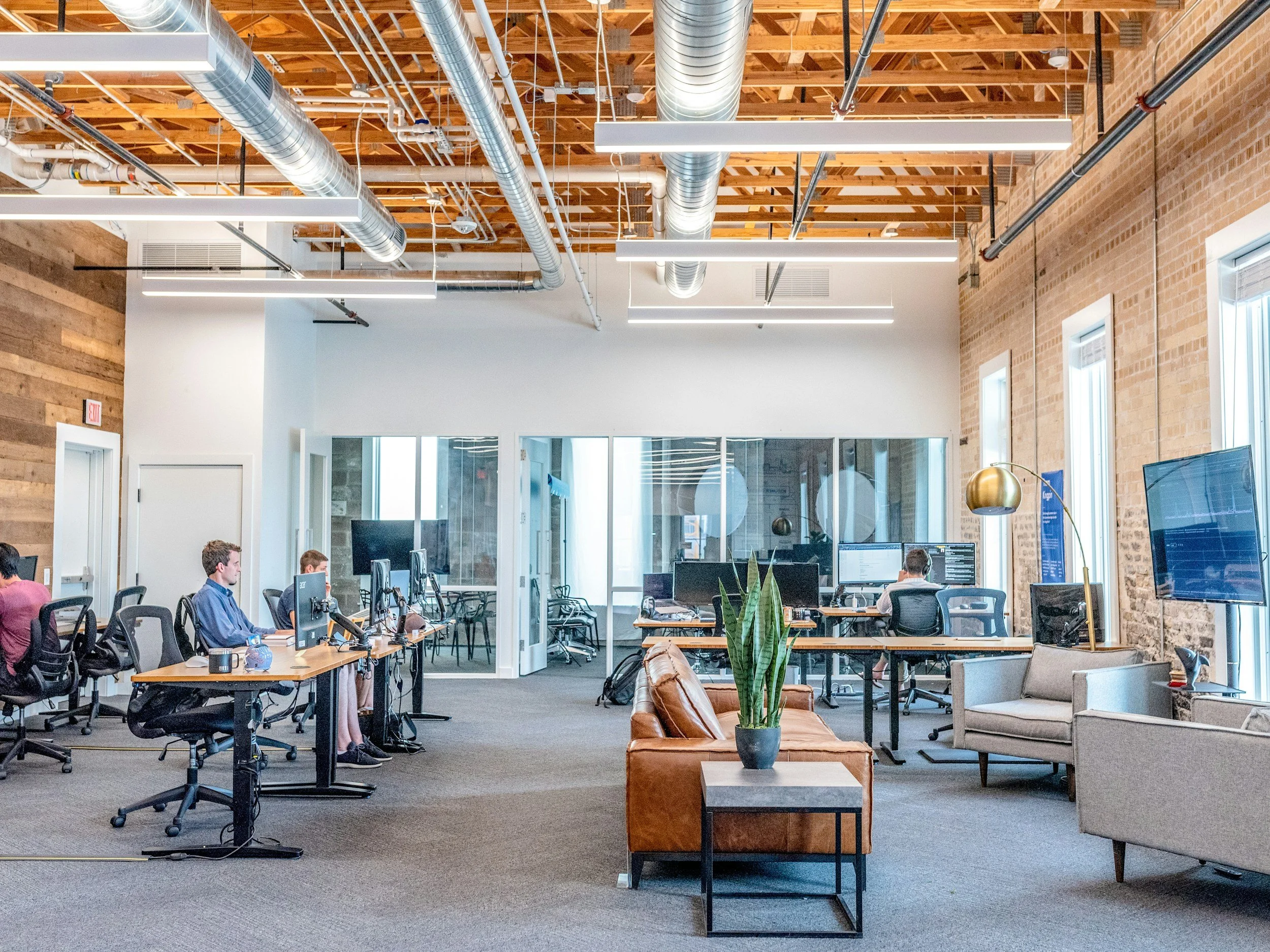 Open-concept modern office with exposed brick walls, large windows, and a mix of workstations and seating areas. Several employees work at desks with computer monitors; there is a glass-walled conference room in the background. The ceiling has exposed ducts and wooden beams, and there are various lighting fixtures including linear LED lights and a gold desk lamp. Interior plants add greenery to the space.