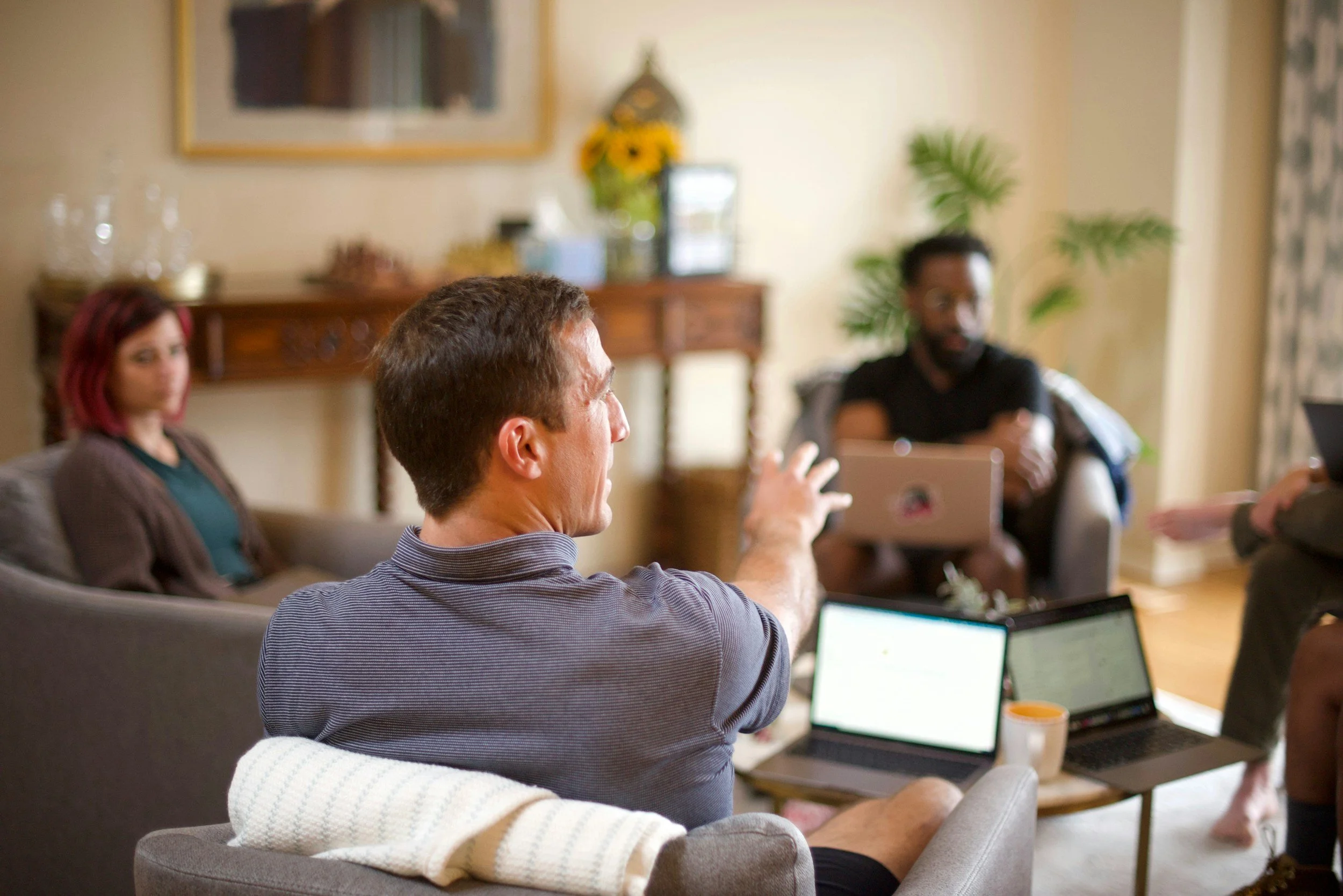 A group of people engaged in a discussion in a cozy living room, with laptops and notebooks on a coffee table.