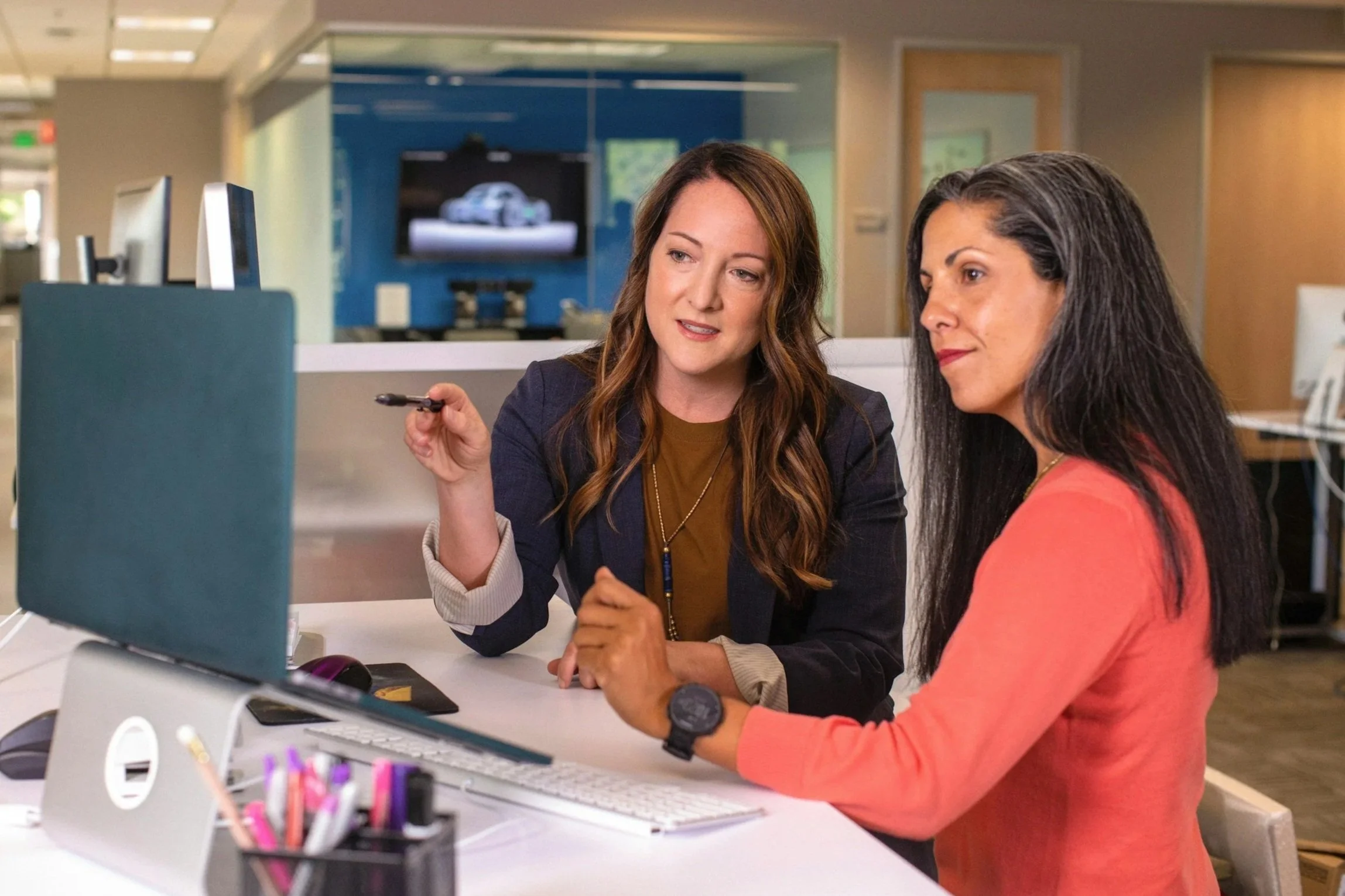 Two women sitting at a desk in an office, looking at a computer monitor and discussing something.